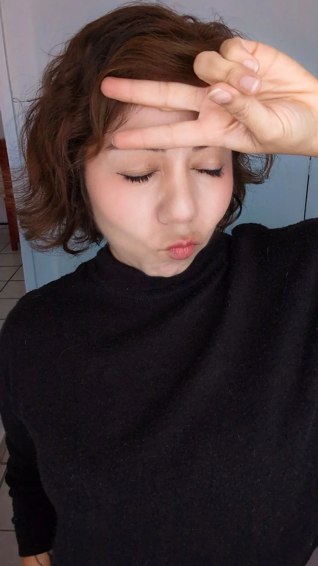 Artist Dharma Dharling: a woman with short, wavy brown hair closes her eyes and makes a kissy face, holding up her right hand in a peace sign near her face, wearing a black turtleneck.