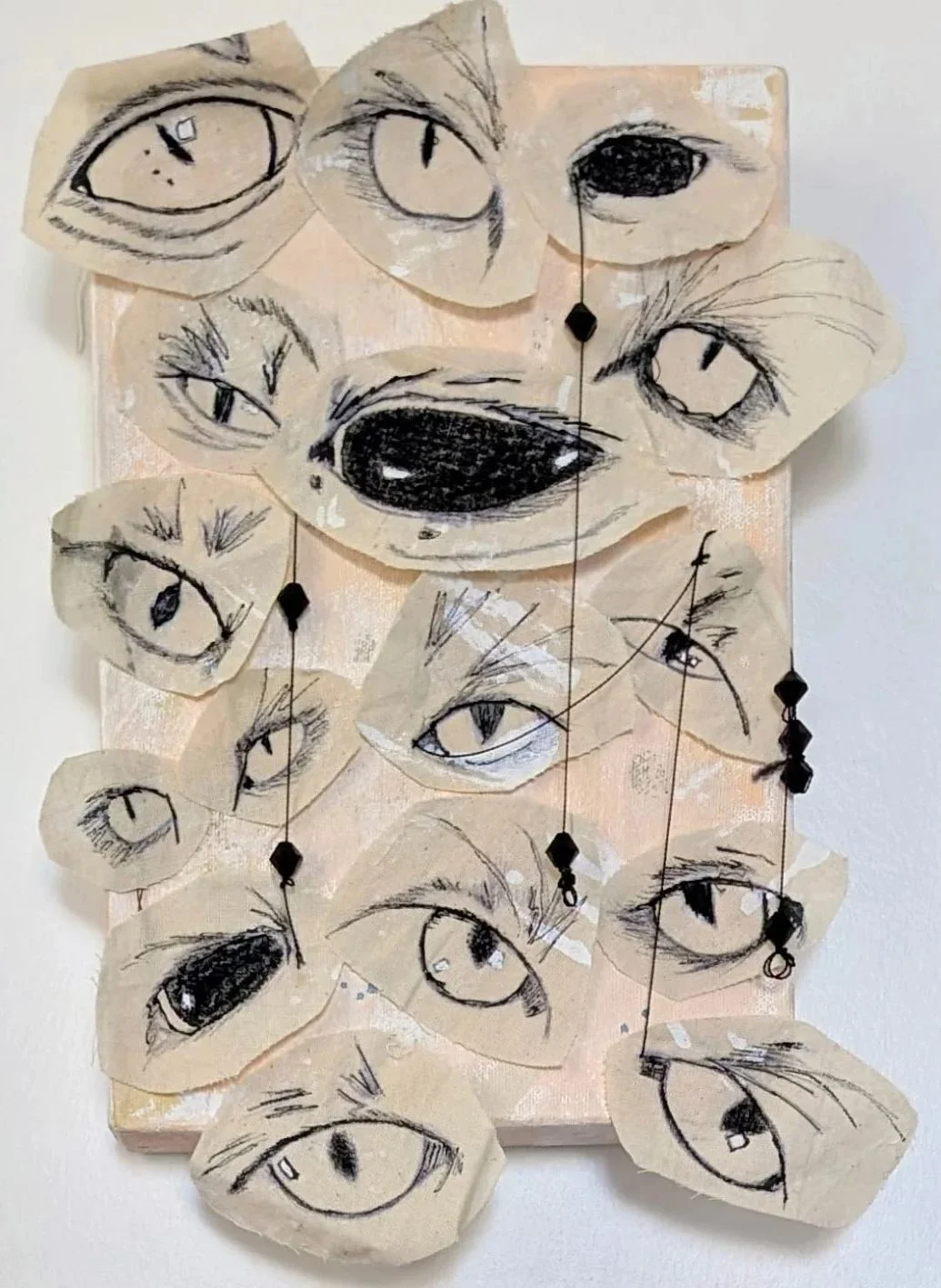 Mixed media artwork featuring multiple sketches of cat eyes on torn pieces of fabric, arranged on a canvas with hanging black beads.