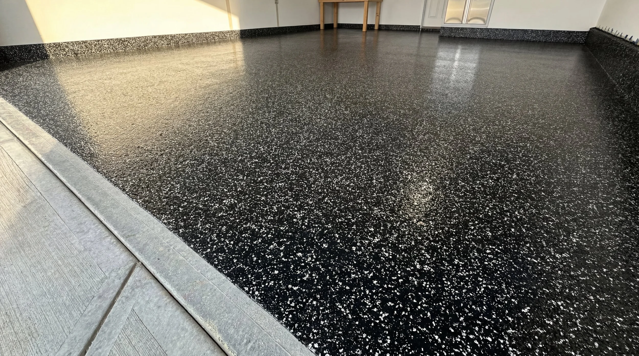 Close-up of a black epoxy resin garage floor with white speckles, a silver metal drain cover at the edge, and part of a wooden table in the background.