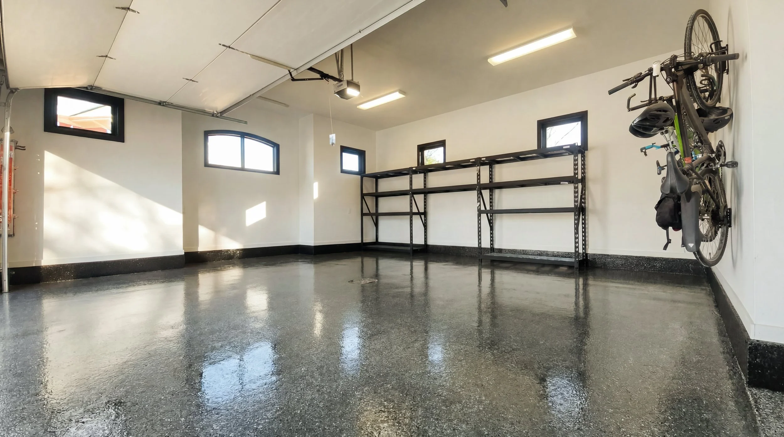 Empty garage with black shelving units, a mounted bicycle with helmets, and small windows on white walls, polished concrete floor, overhead lights