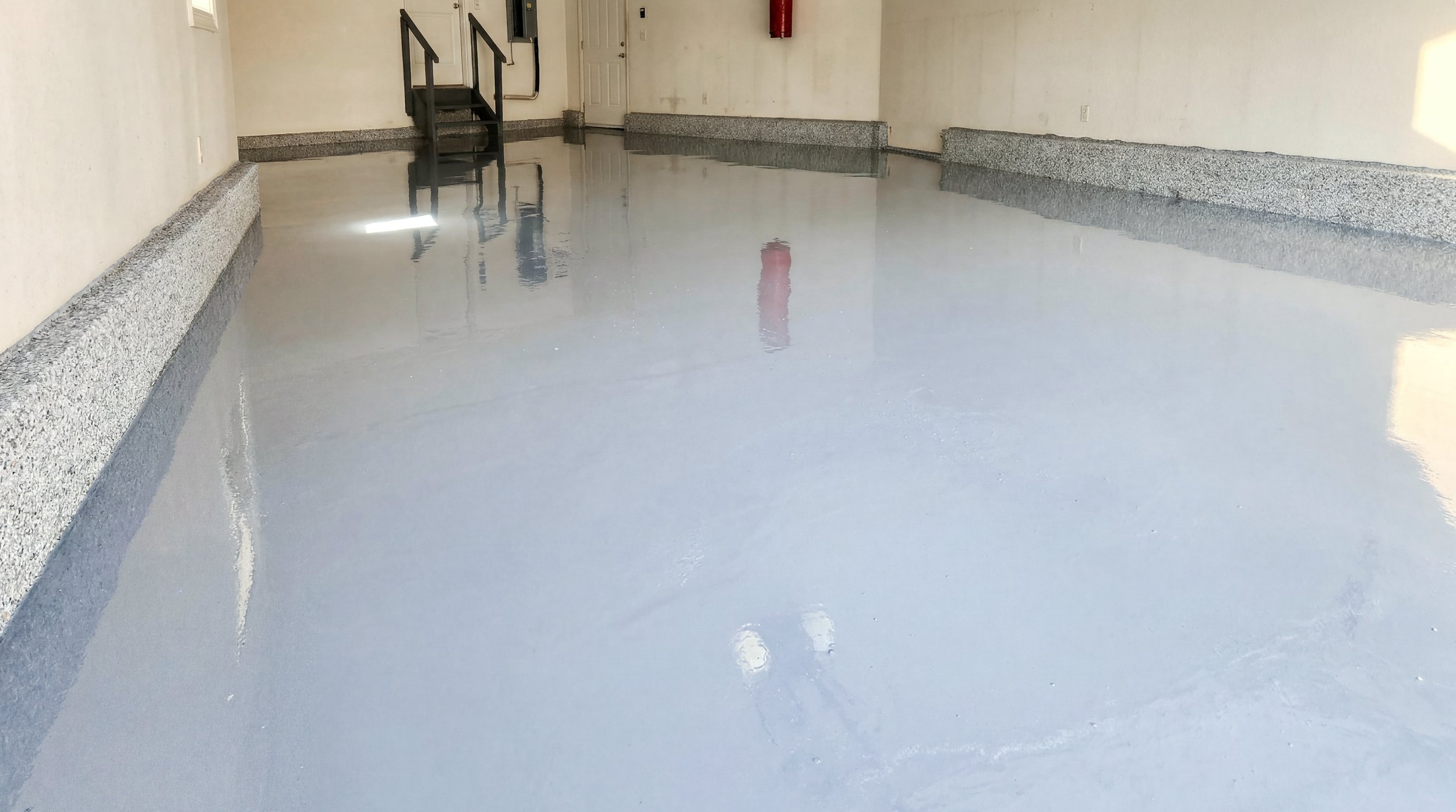 Room with a shiny, gray epoxy floor, white walls, a staircase with metal railings, a door, and a fire extinguisher mounted on the wall.