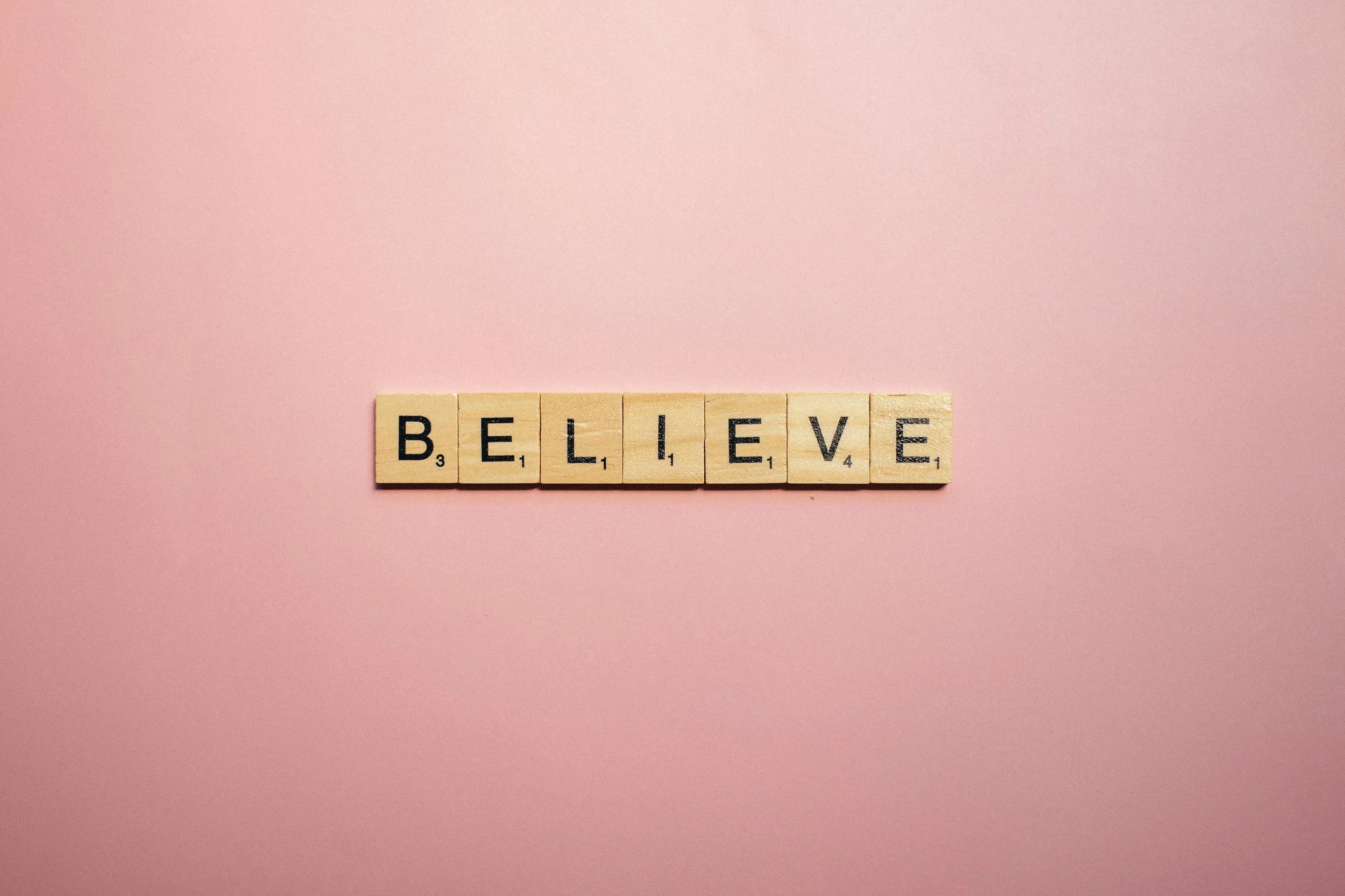 Wooden Scrabble tiles spelling out 'BELIEVE' on a pink background.