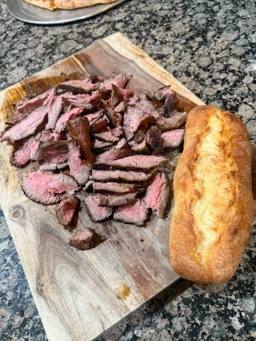 Sliced cooked steak on a wooden cutting board with a loaf of bread on the side.
