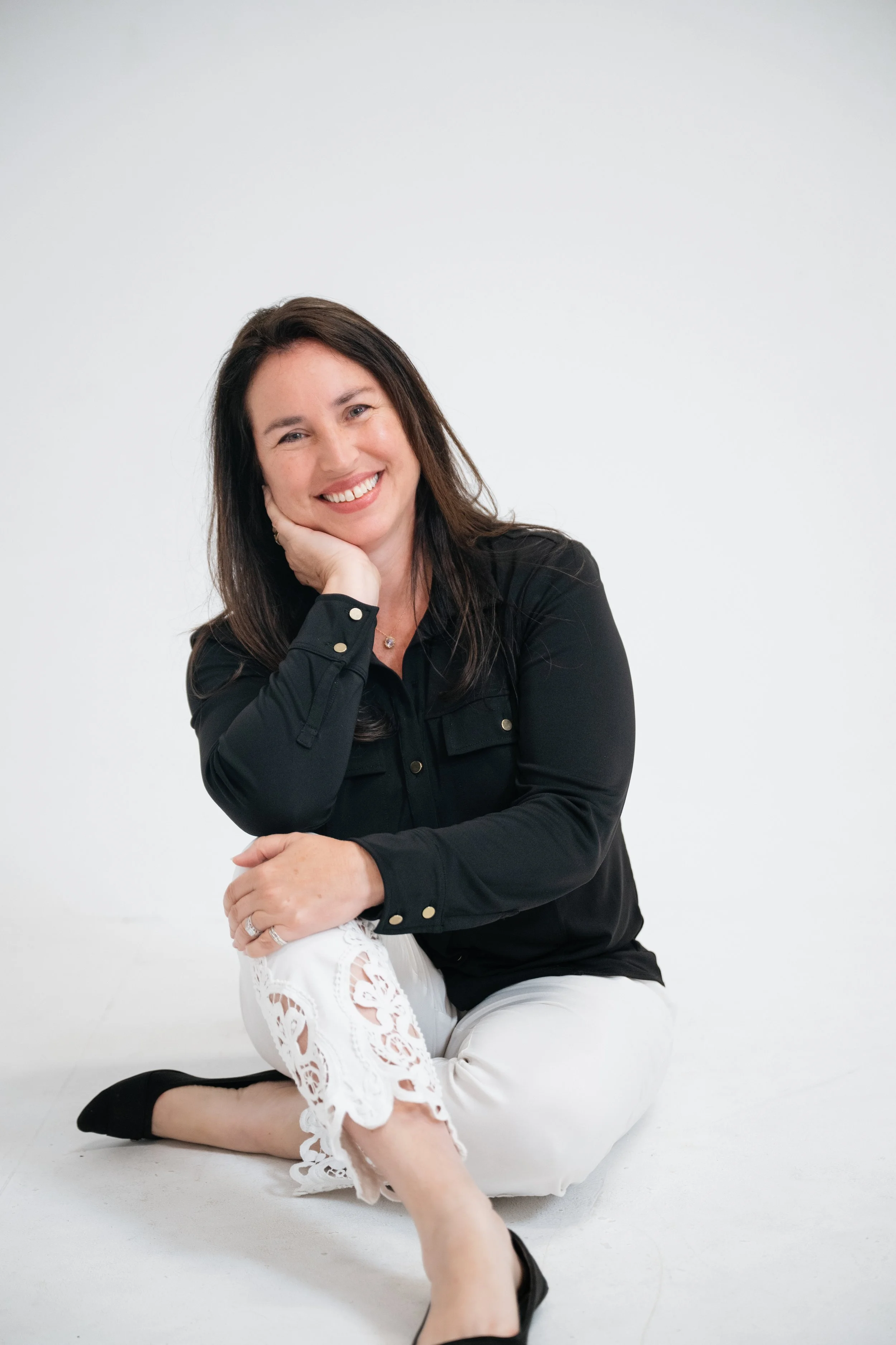 A woman smiling, sitting on the floor with one knee bent and resting her chin on her hand, dressed in black top and white lace pants, against a plain white background.
