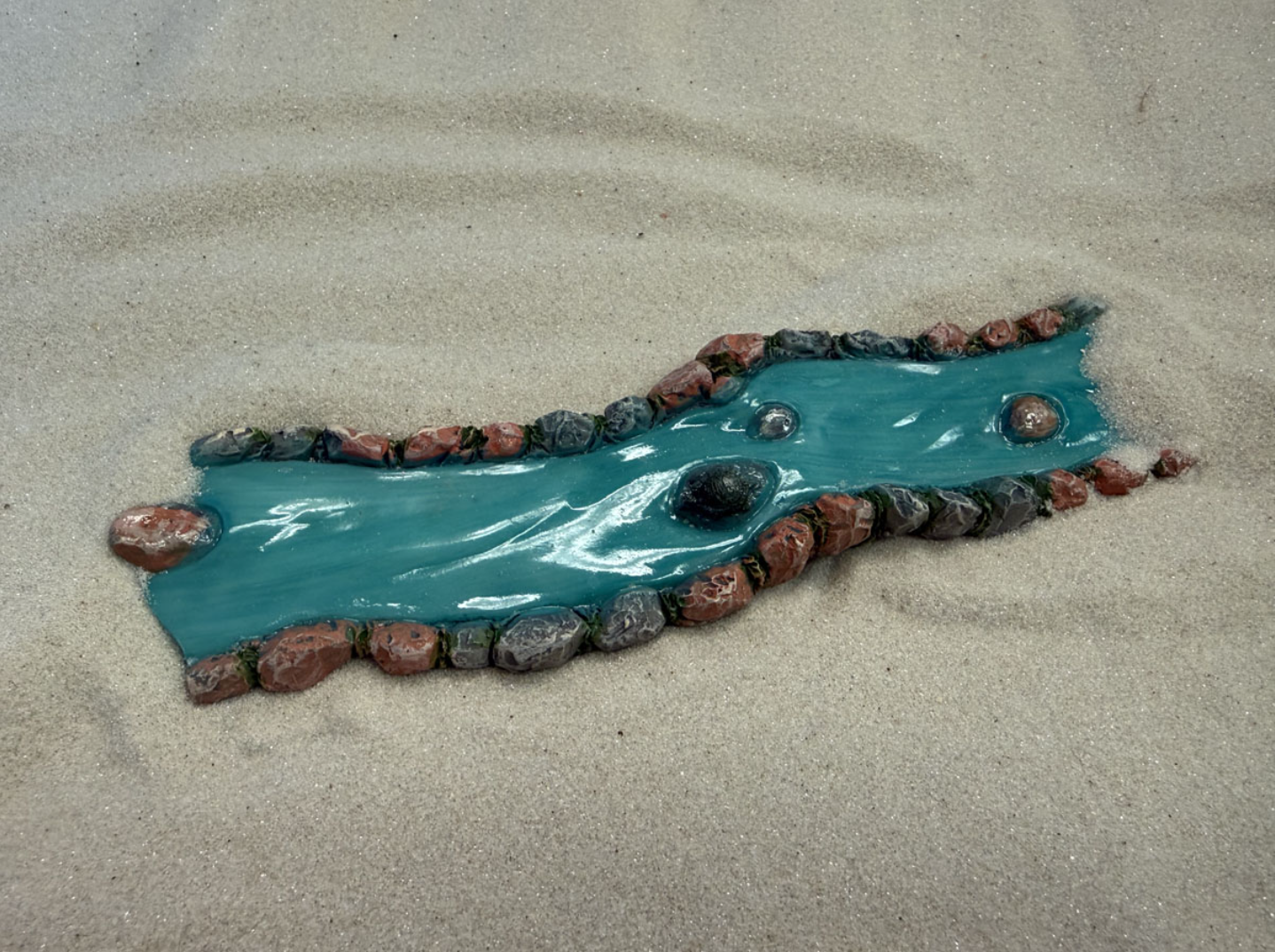 A small turquoise plastic toy crocodile with its body outlined by a border of multicolored rocks on sandy ground.