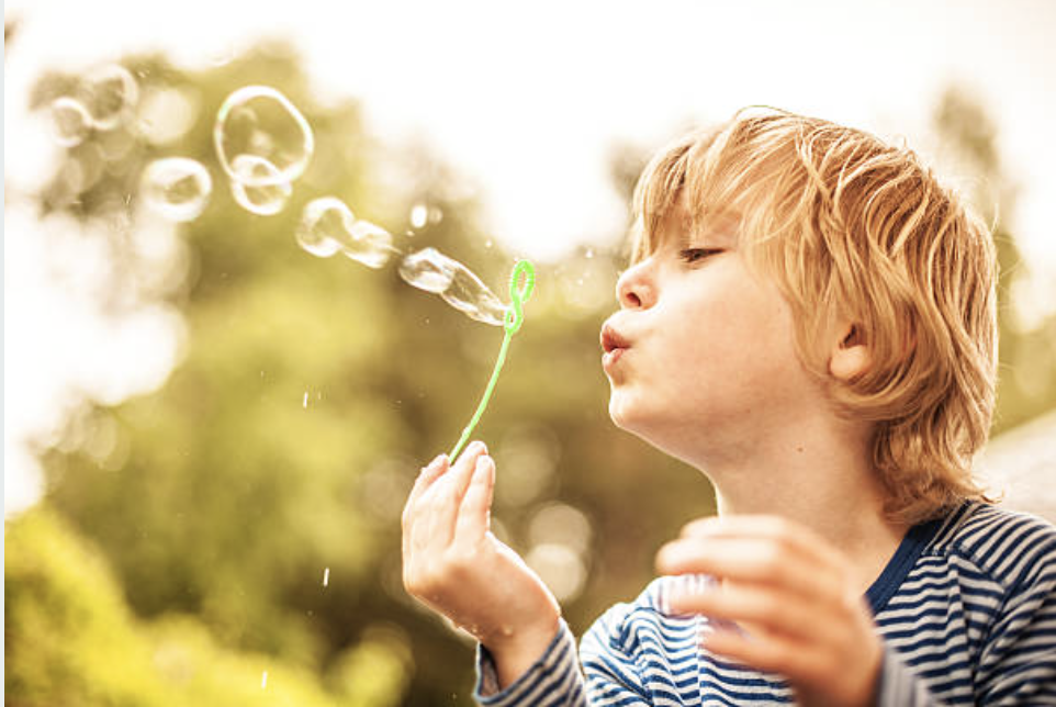 A young boy with red hair blowing bubbles outdoors on a sunny day.