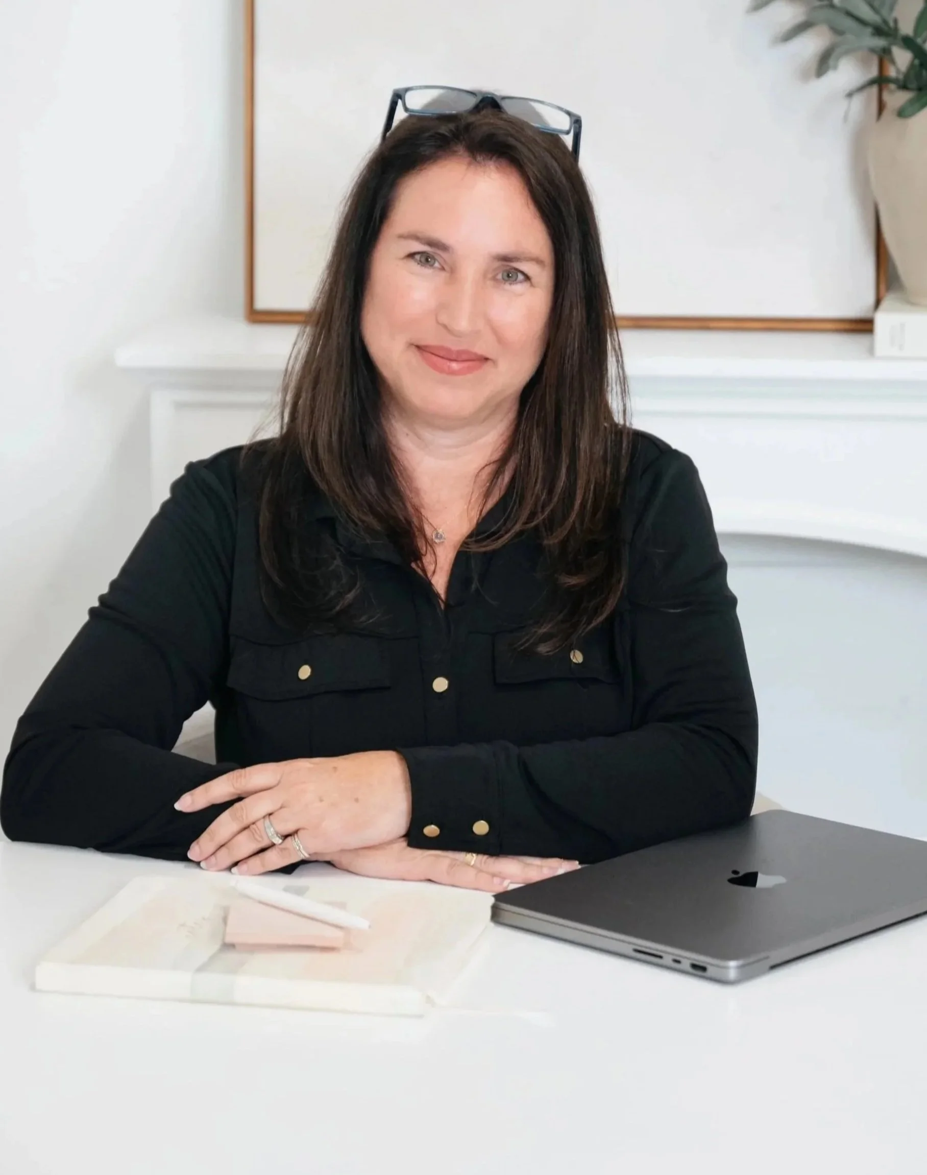 A woman with long dark hair and glasses on her head, wearing a black top, sitting at a white desk with a closed laptop, a white notepad, and some papers, in a bright room with a potted plant in the background.