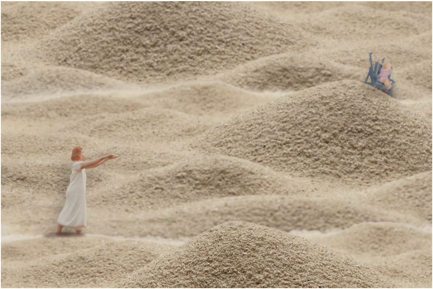 Miniature figurines of three people in a desert or sandy environment. One woman in a white dress is standing with outstretched arms, while two other figures are sitting on a bench or chair in the distance.