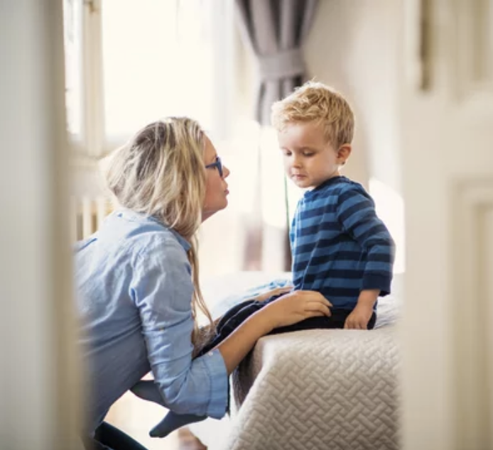A woman and a young boy in a bedroom, engaging in a close conversation. The woman is sitting on the bed, leaning forward towards the boy, who is standing on a surface. The room has natural light, curtains, and a neutral color scheme.