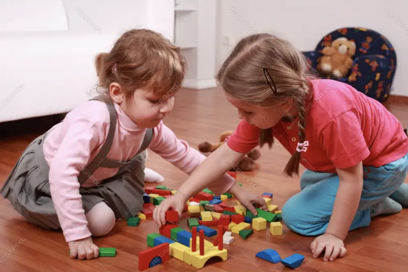Two young girls playing with colorful building blocks on a wooden floor indoors.
