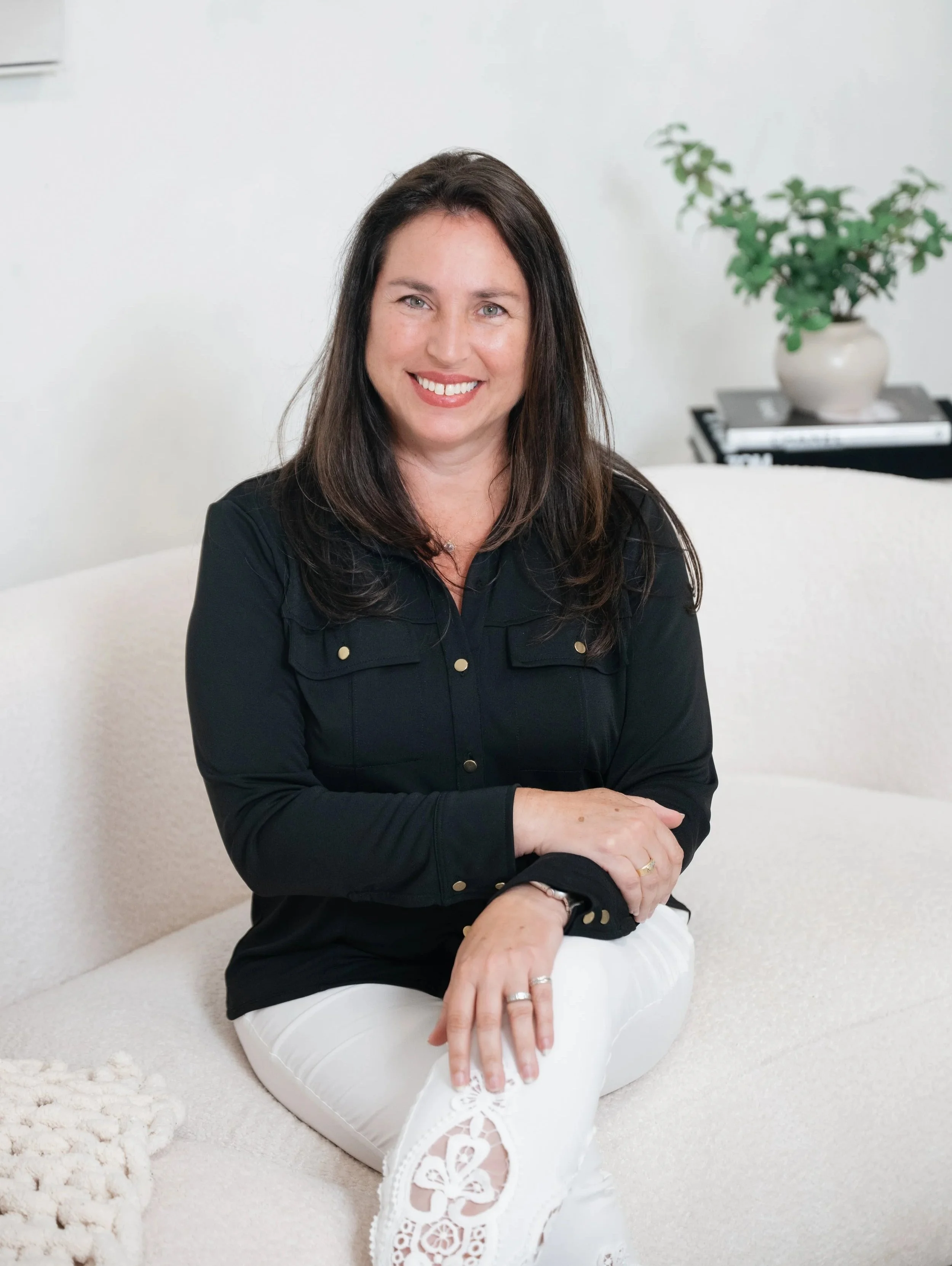 A woman with long brown hair and a bright smile sitting on a white sofa with a white cushion. She is wearing a black jacket and white pants with lace details. There is a potted plant in the background on a table.