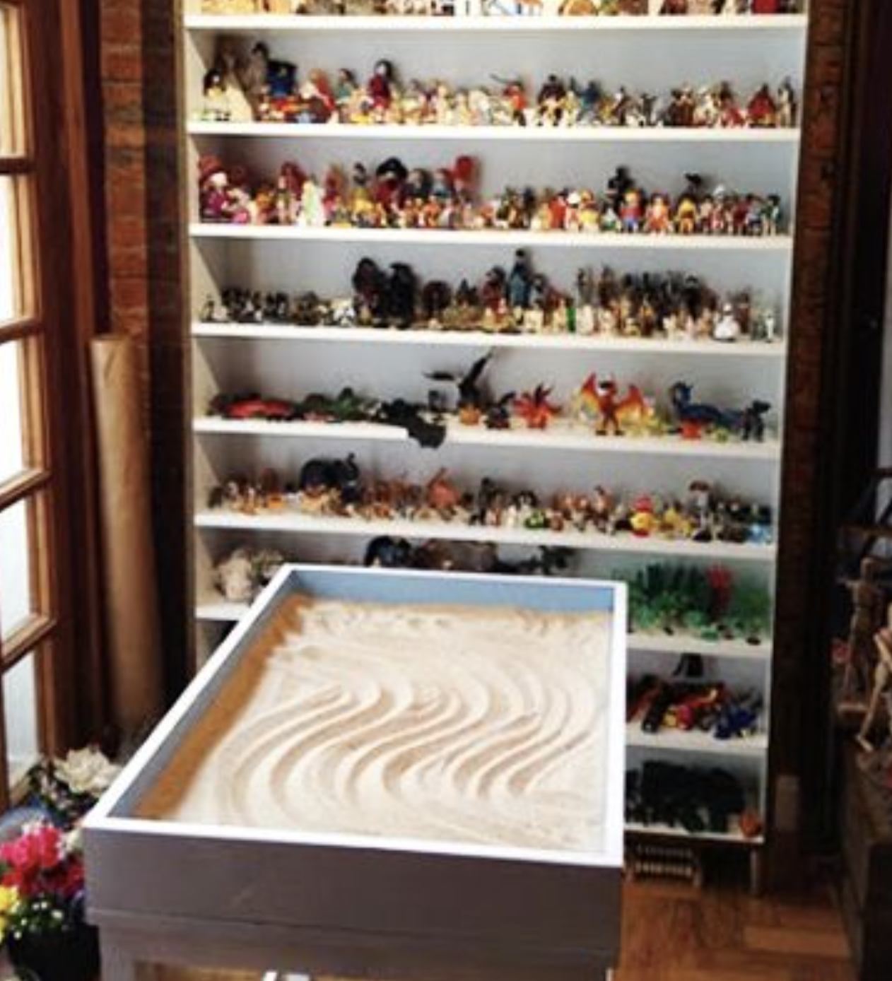 Shelves filled with small, colorful toy figurines and models, and a table with a sand art display in front of the shelves.