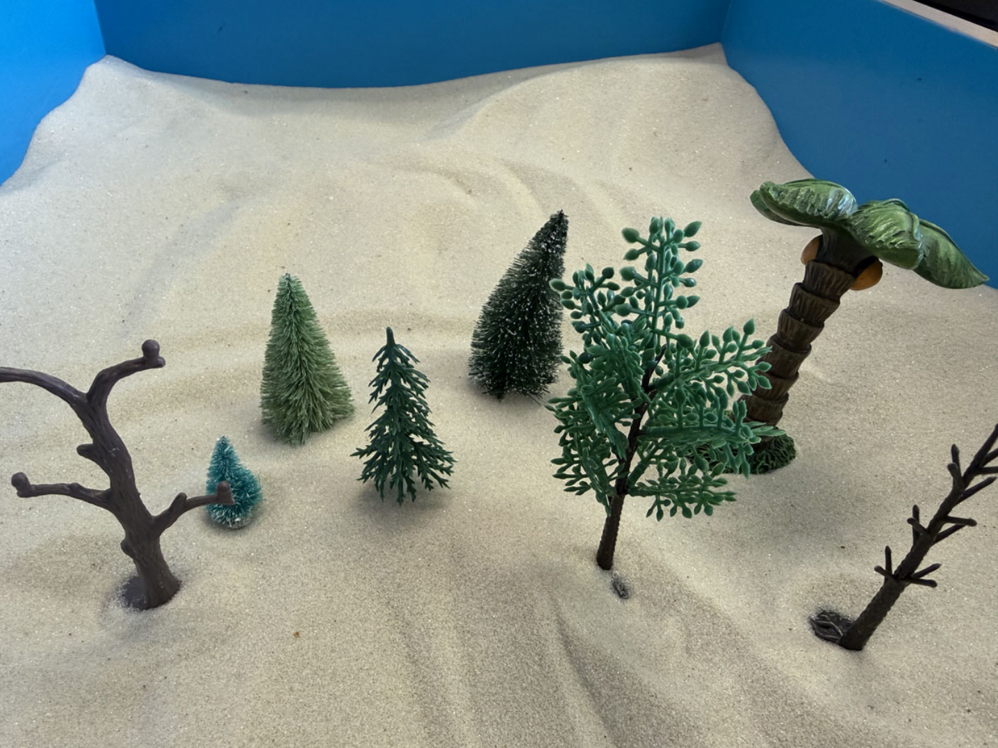 Miniature model of a desert landscape with small plastic trees and a fake palm tree on light-colored sandy ground.