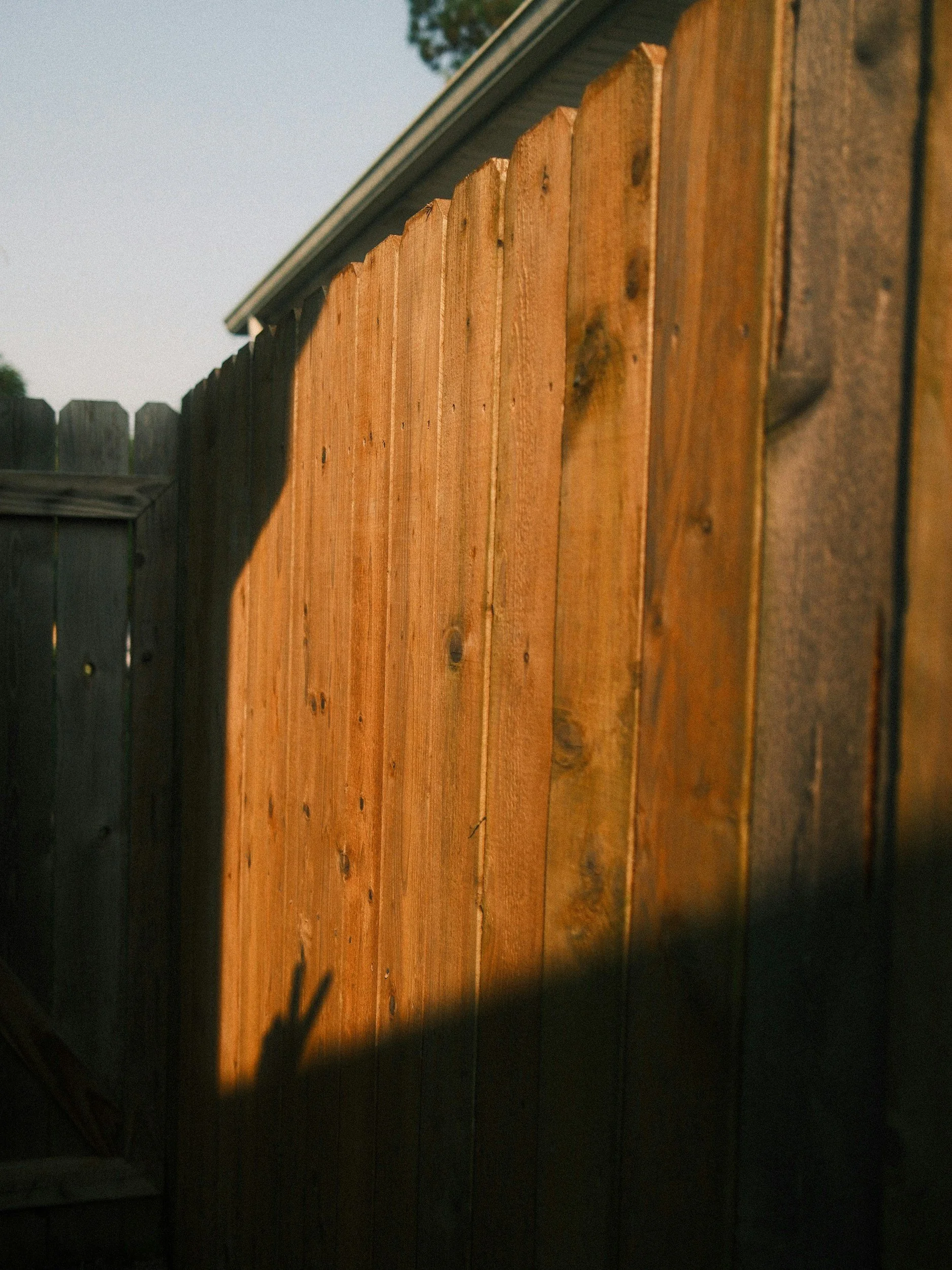 Close-up of a wooden fence with a dark shaded area at the bottom and a house roof in the background under a clear sky.