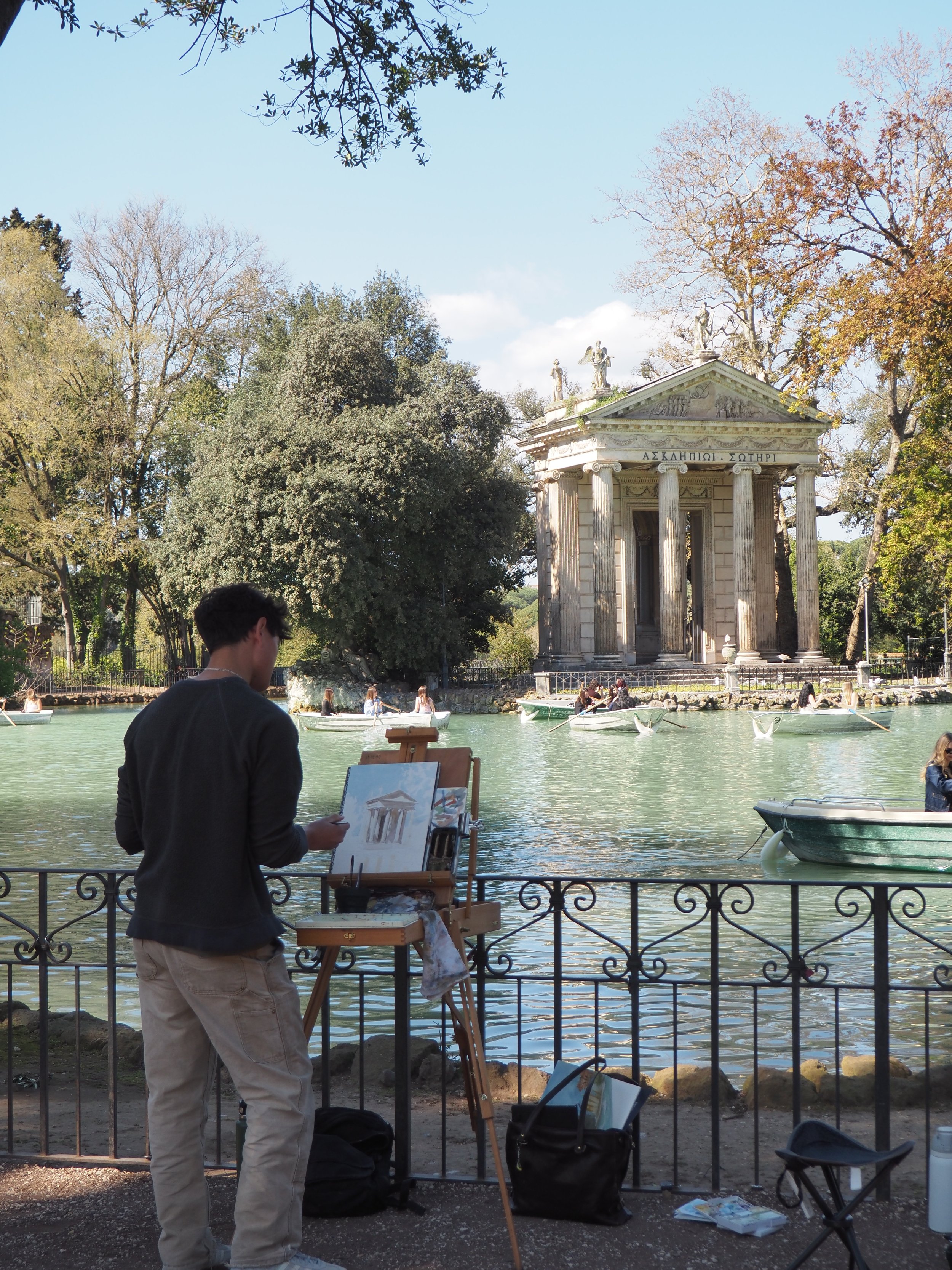 Artist painting the Temple of Aesculapius beside Rome’s Villa Borghese lake, where visitors row boats among the trees.
