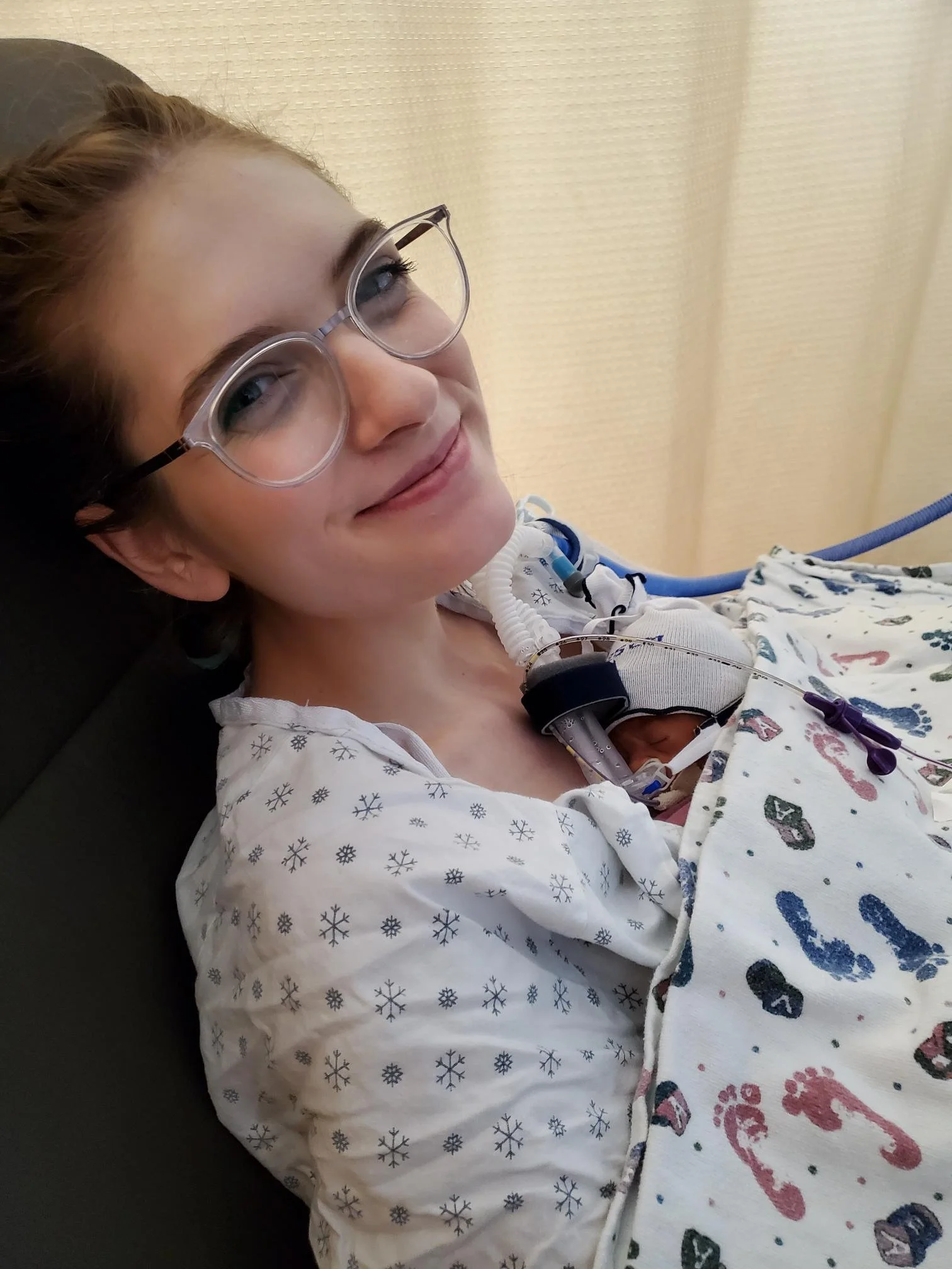 Young woman with glasses lying in a hospital bed, connected to medical tubing and wearing a hospital gown with Christmas-themed print.