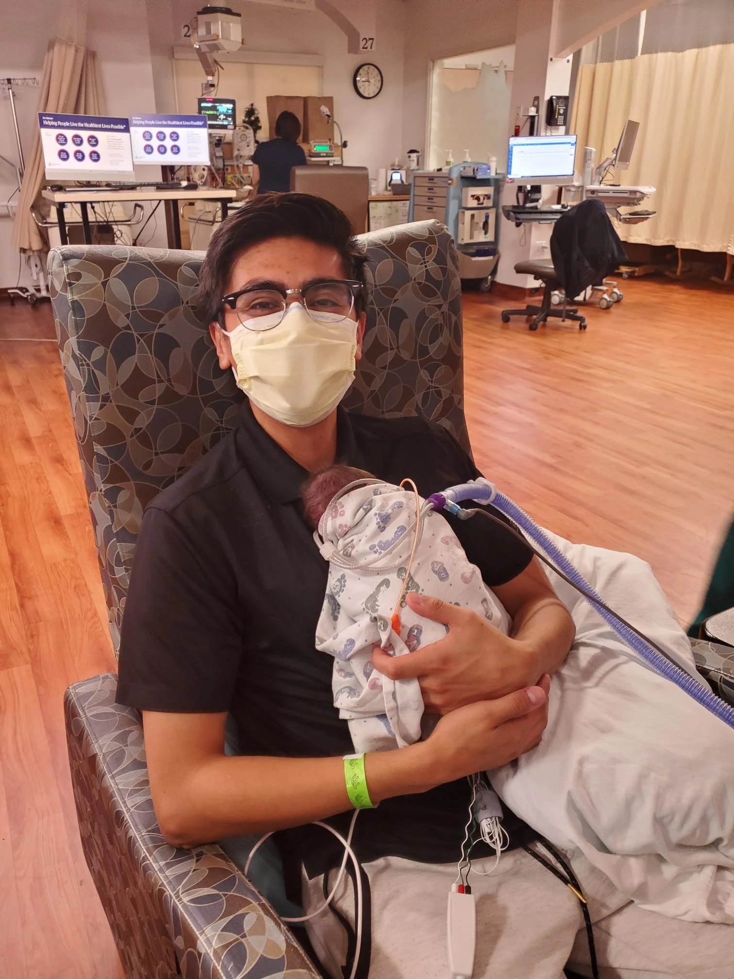 A young man wearing glasses and a face mask is sitting in a hospital room holding a newborn baby, who is connected to medical tubes and monitors. The room is equipped with medical equipment, monitors, and monitors showing medical data.