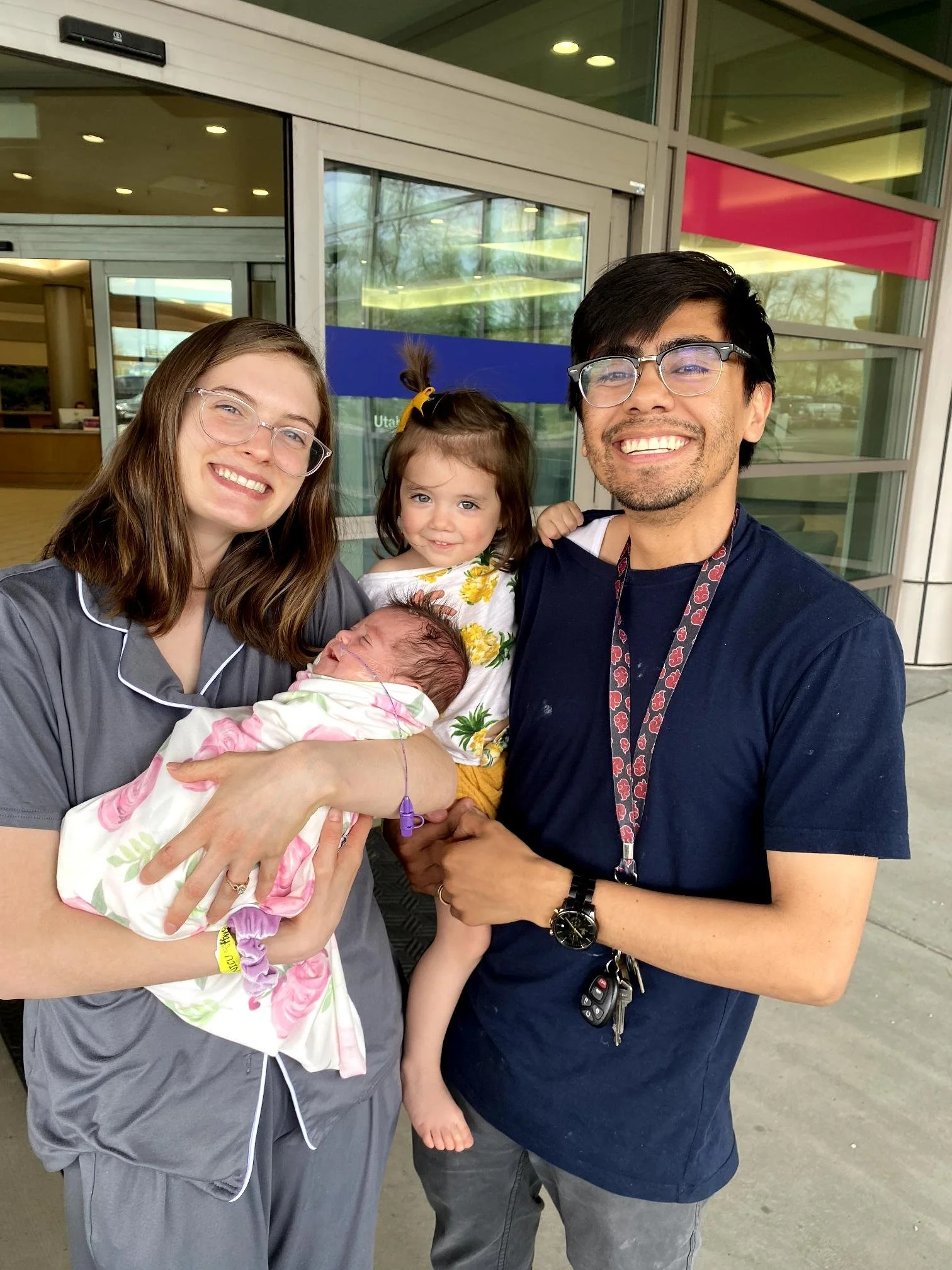 A group of four people standing outside a modern building, smiling for the photo. The group includes women, a girl, and a man holding a newborn baby. The woman on the left is wearing glasses and a gray outfit, the girl has curly hair with a yellow bow and is dressed in a floral outfit, and the man is wearing glasses, a navy blue shirt, and has a set of keys hanging from a lanyard around his neck.