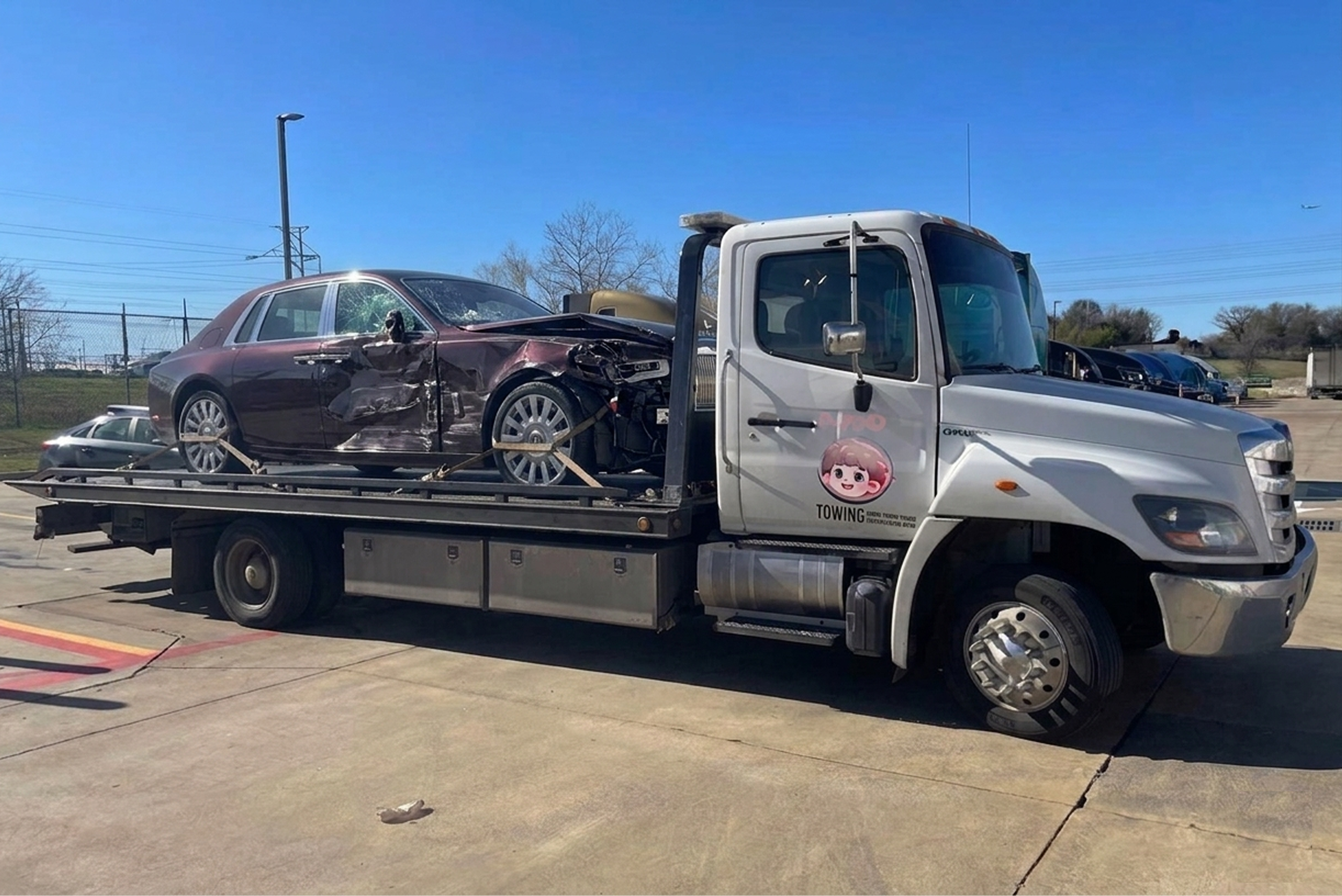 A white tow truck carrying a damaged red car with a shattered windshield on a parking lot under a clear blue sky.