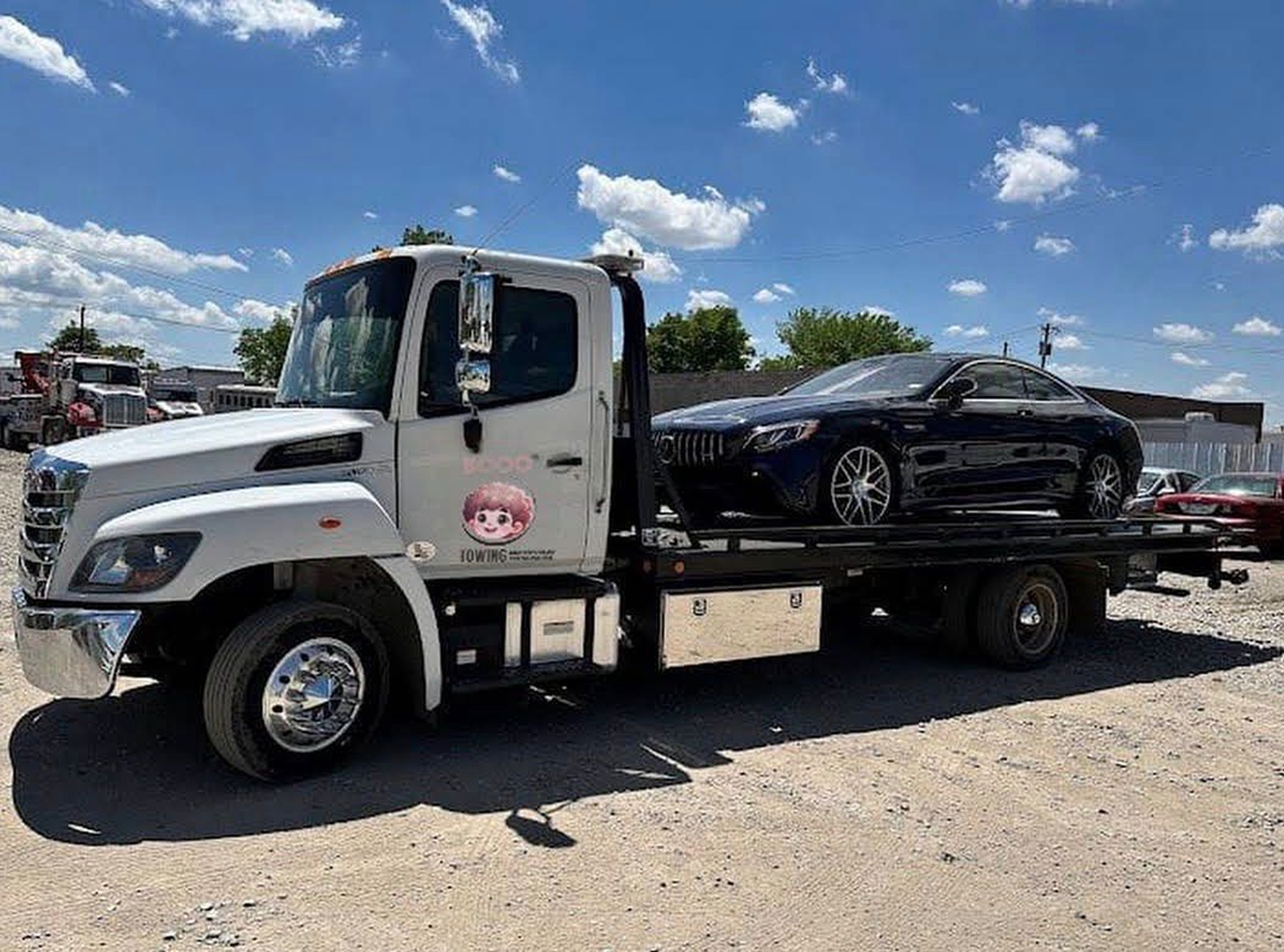 A flatbed tow truck is carrying a black sports car on its bed, parked on a gravel lot under a partly cloudy blue sky with green trees and other parked trucks in the background.