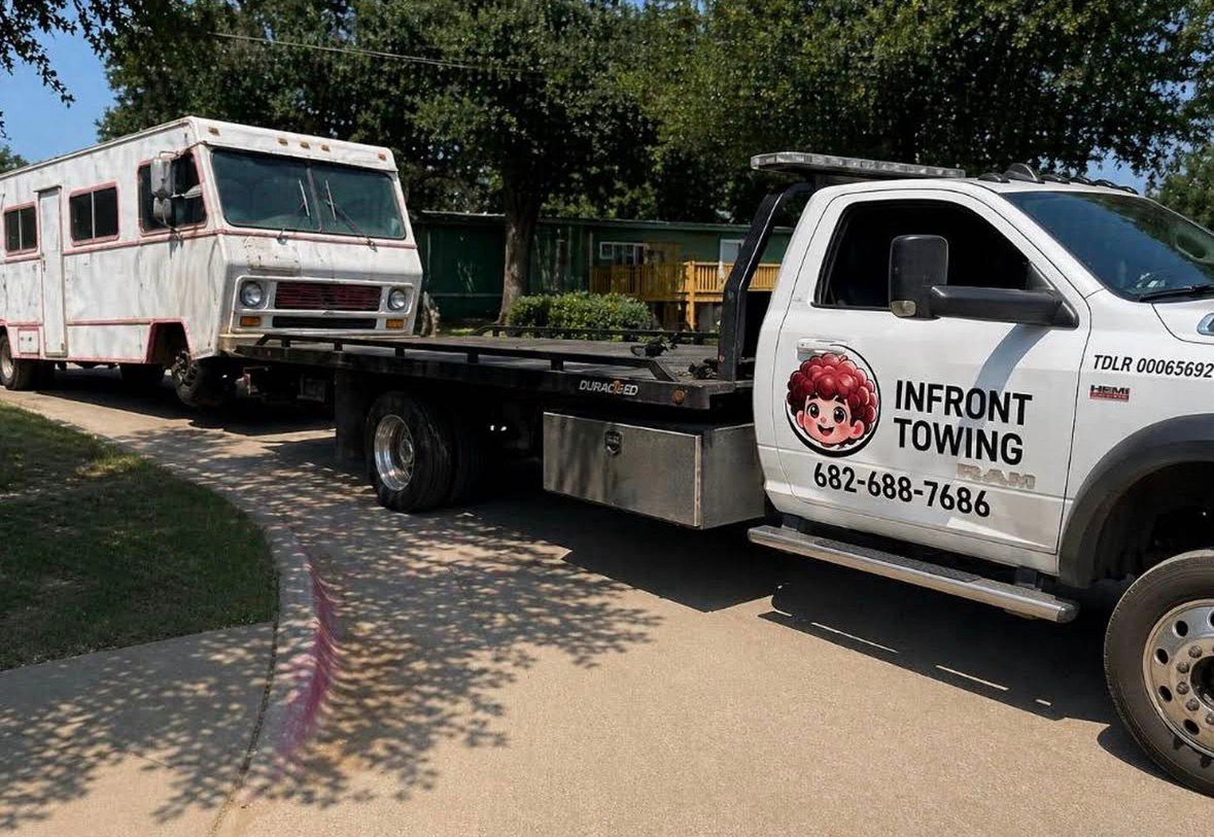 A white tow truck with a cartoon character's face on the door, towing an old, rusty RV on a flatbed trailer.