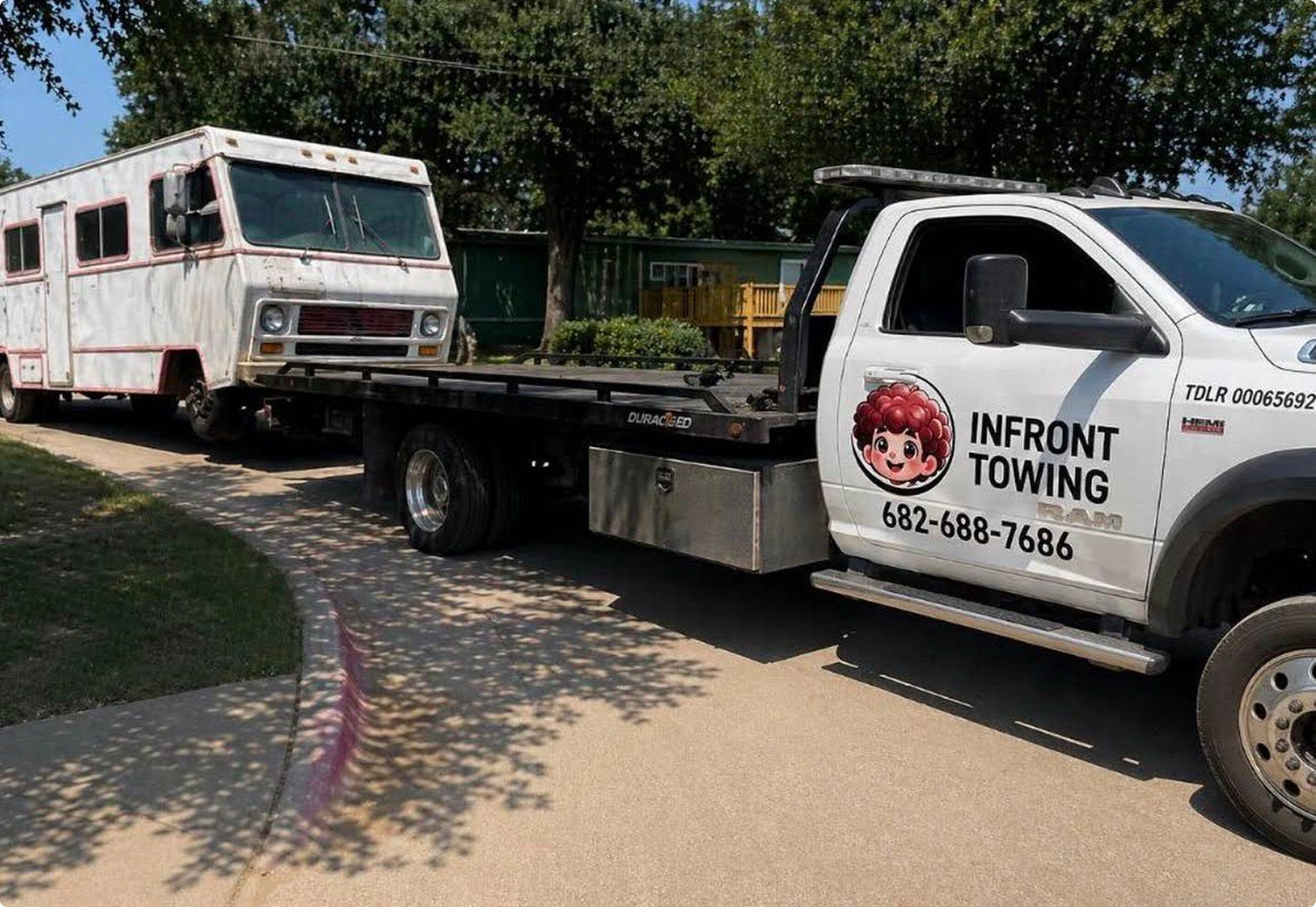 A tow truck with the logo and name 'InFront Towing' is towing an old, white camper trailer on a street with trees and houses in the background.