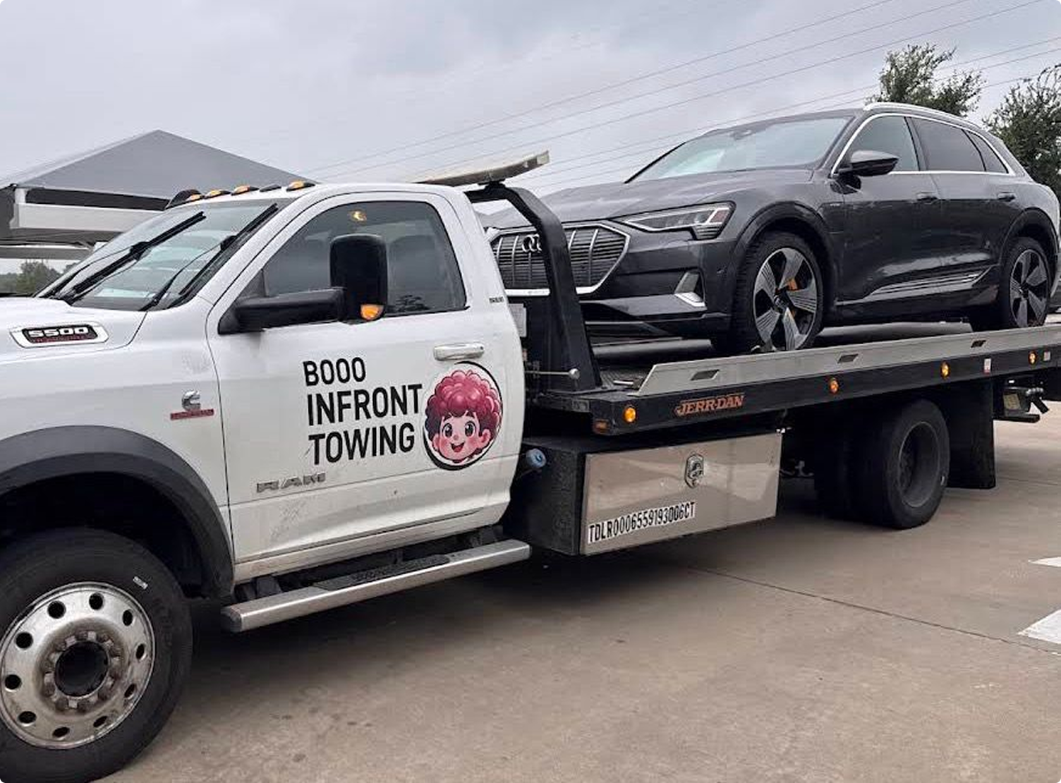 A flatbed tow truck carrying a black Audi car, with the truck displaying the text 'BOO IN FRONT TOWING' and a cartoon girl's face on the door.