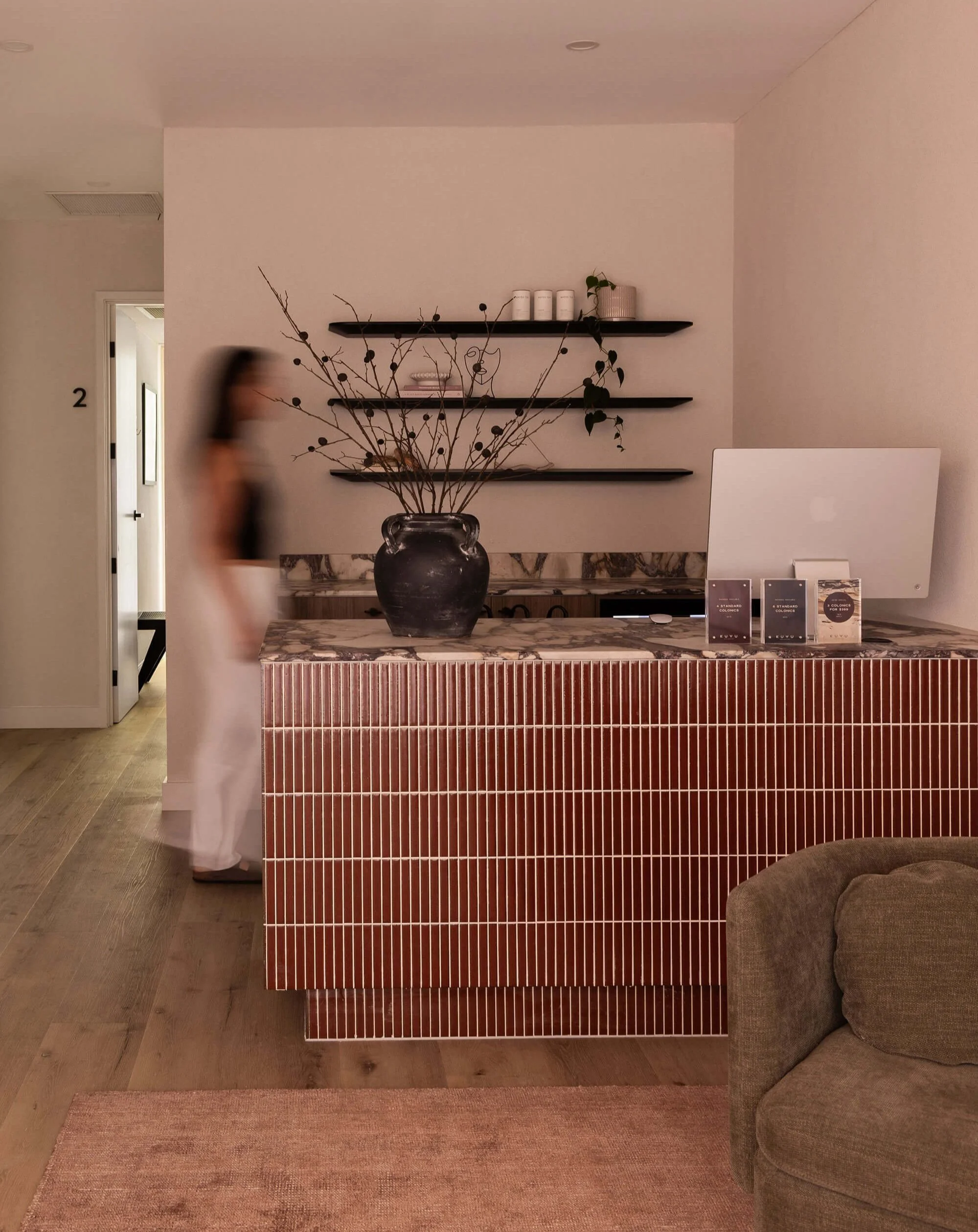 Interior of a modern room with a marble-topped counter, a large black vase with dried branch and seed pods, black wall-mounted shelves with decorative items, a door with the number 2, a woman's blurred figure walking past, a computer on the counter, and a brown upholstered chair in the foreground.