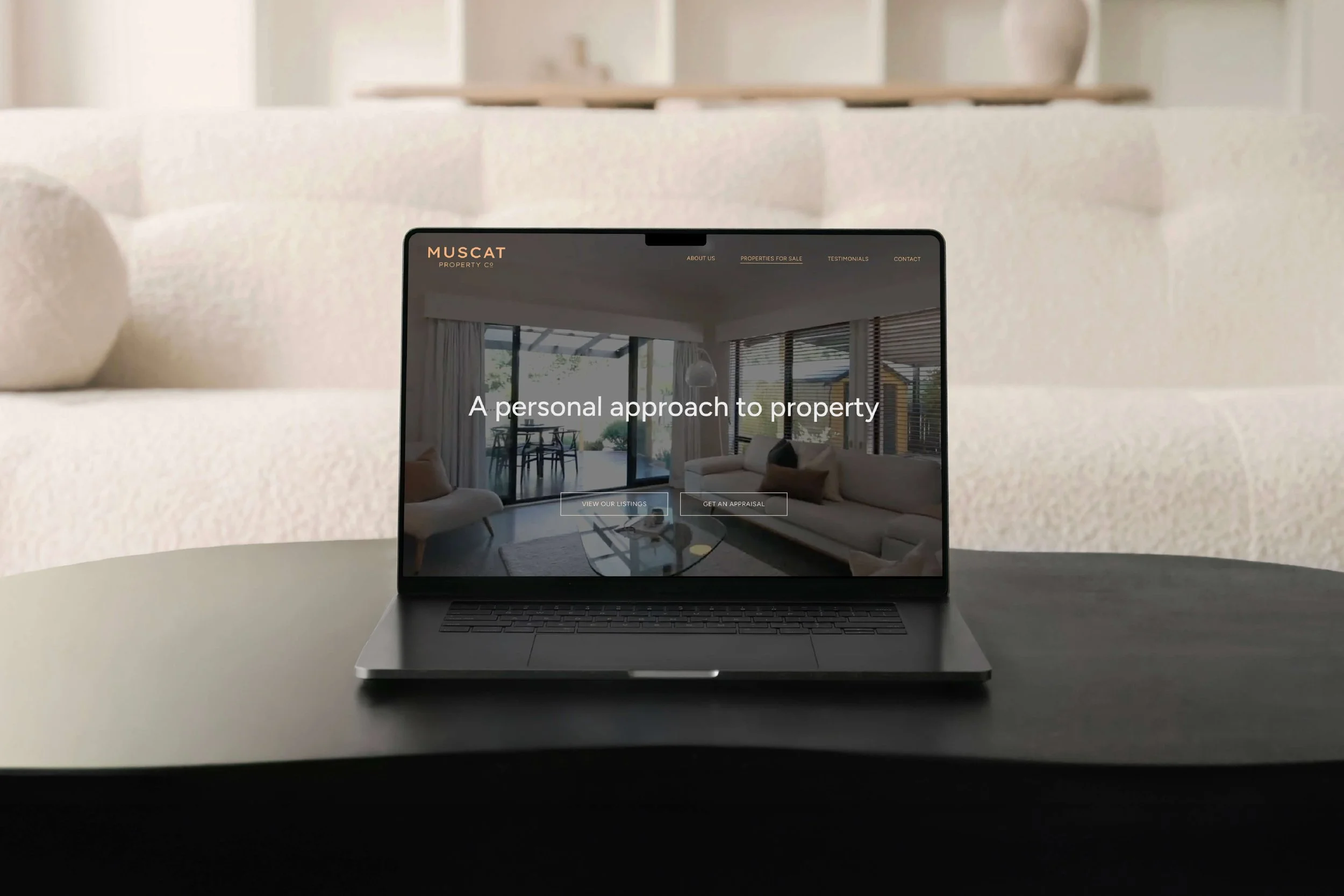 A laptop on a white table displaying a real estate website called Muscat Property Co, with a background of a modern, well-lit living room with large windows, a sofa, and a glass door leading outside.