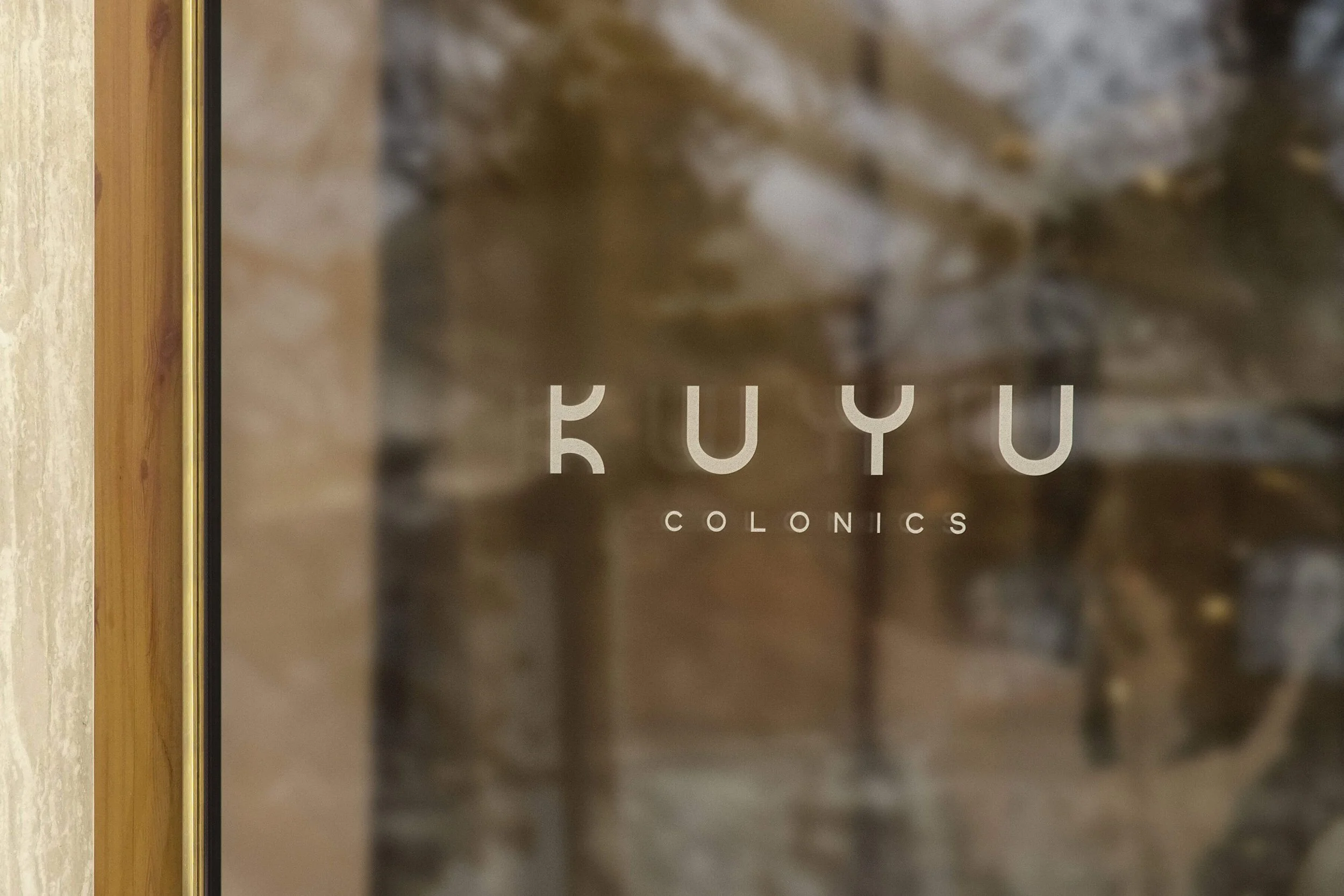 Close-up of a door or window with a glass panel, showing the words 'Fuyu Colonics' printed on it, with a blurred background of a brick wall.