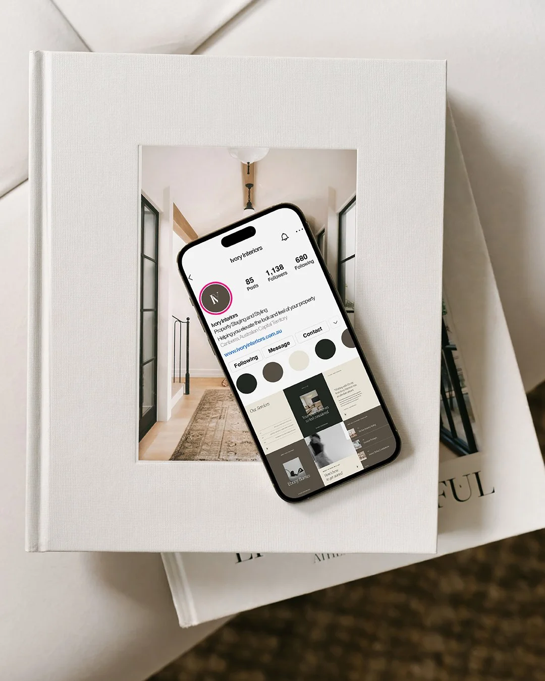 A white hardcover book with a cut-out window displaying a photograph of a modern hallway, placed on top of another white book. An iPhone with a social media profile page is resting on the book, partially covering the photograph.