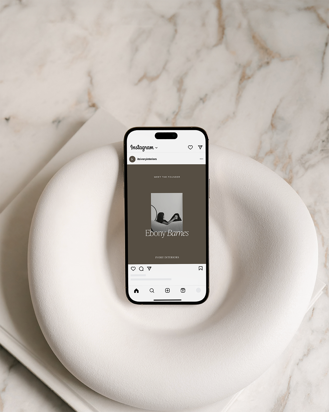 Smartphone displaying an Instagram profile for Ebony Barnes, a founder of Ivory Interiors, placed on a white rounded surface with a marble background.