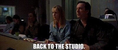 A group of people sitting at a table in a studio or classroom setting, with one person speaking. Text overlay reads "BACK TO THE STUDIO."