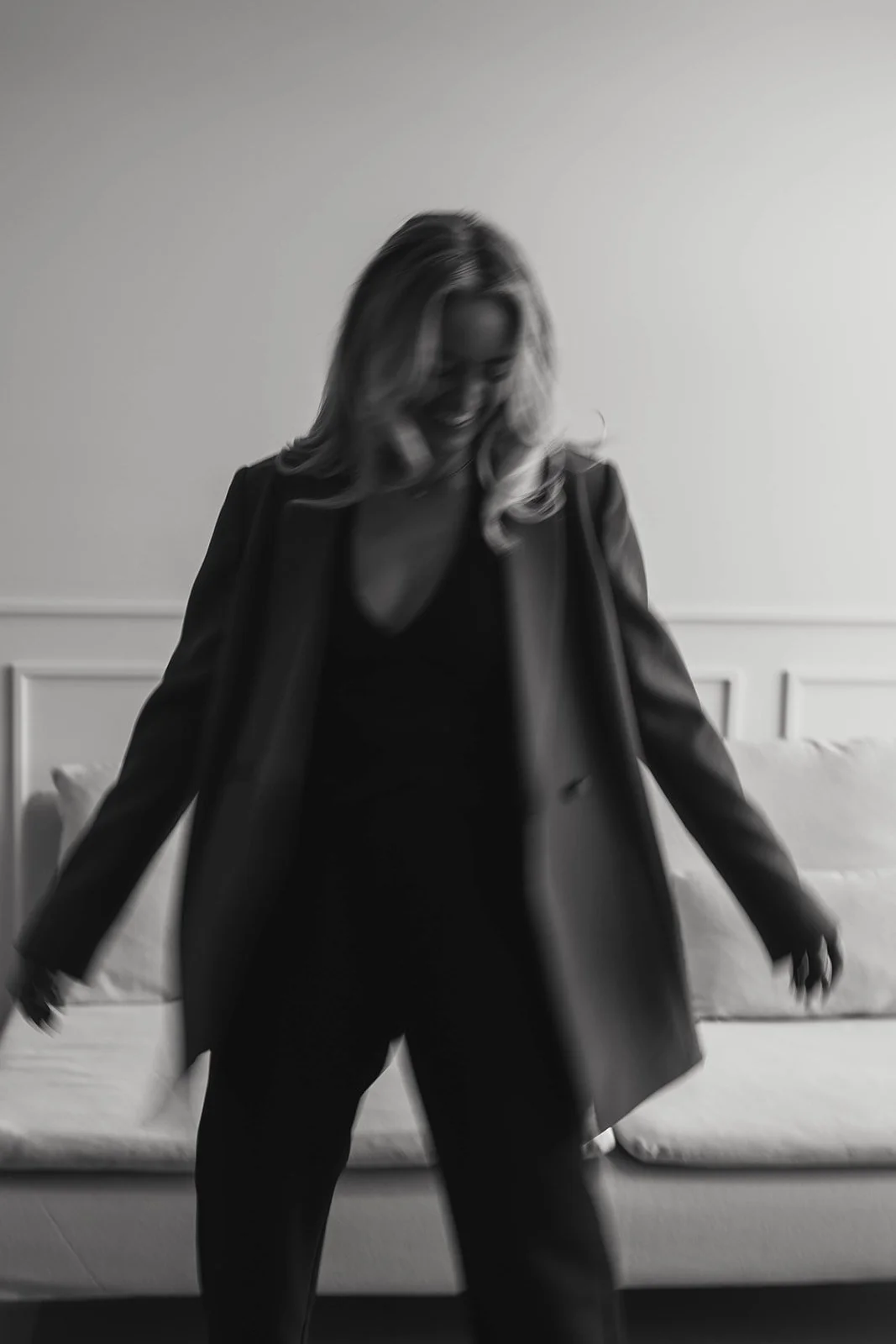 A woman with blonde hair smiling and looking down, wearing a black outfit and an open blazer, standing in front of a bed with a white headboard in a well-lit room.