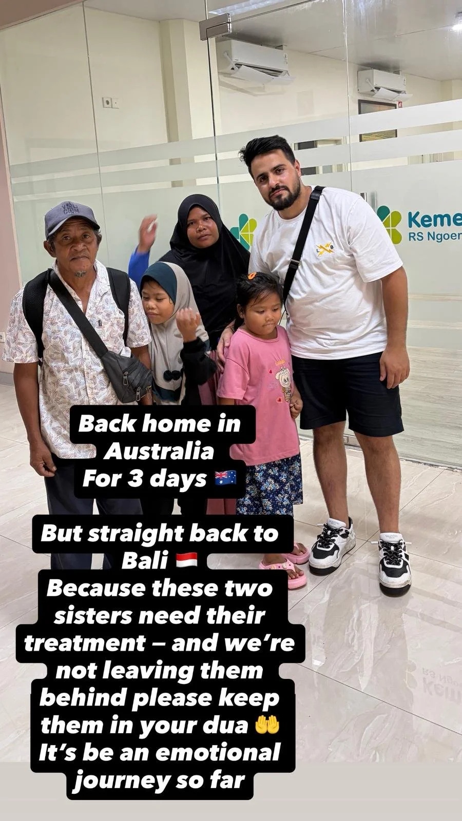 A diverse group of five people, including two children, standing inside a hospital or clinic with a glass wall and medical signage in the background. The group appears to be in an emotional moment following medical treatment in Australia, with some of them looking at the camera.
