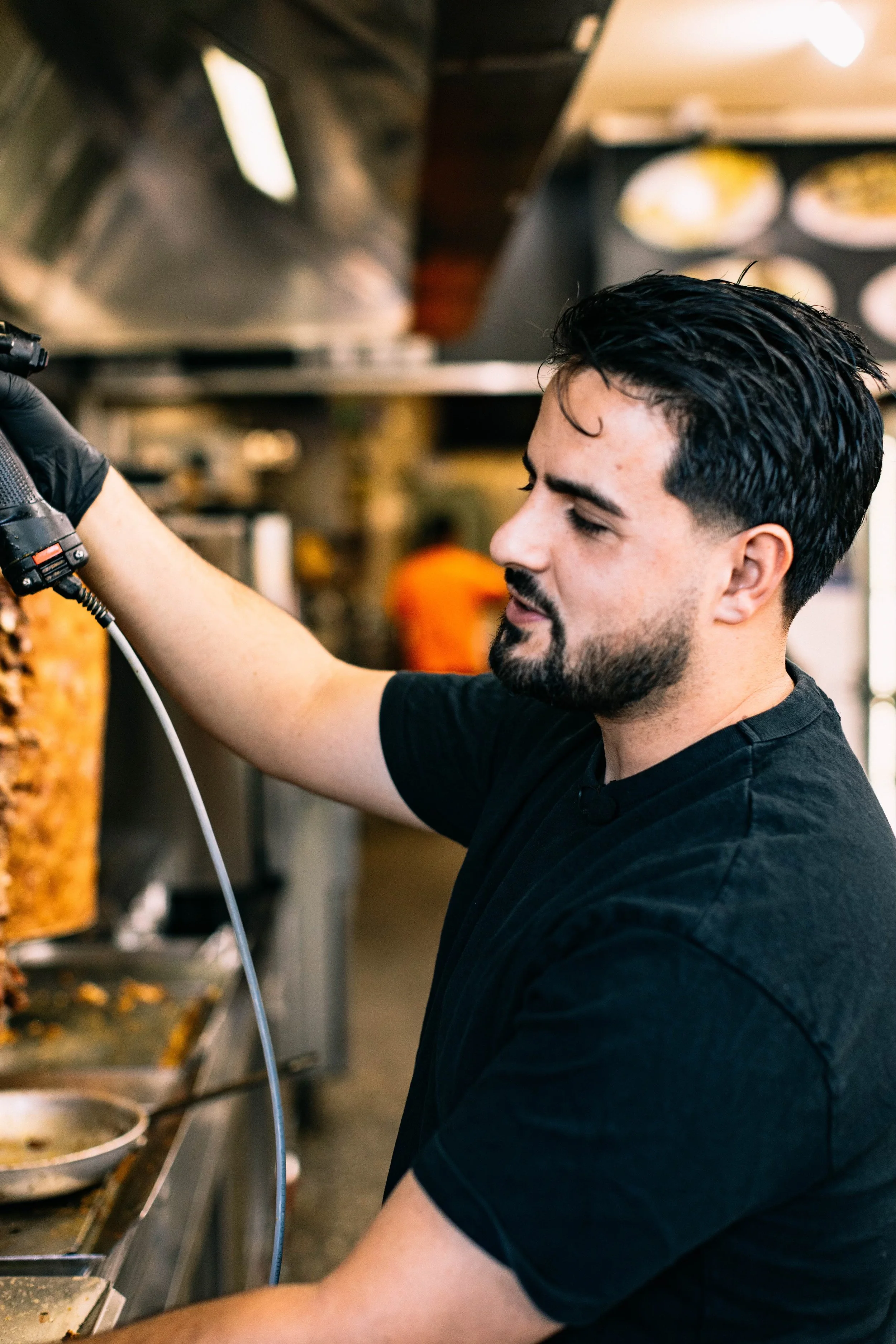 A man with dark hair and a beard wearing a black T-shirt is working in a commercial kitchen, preparing food next to a large grill or stove.