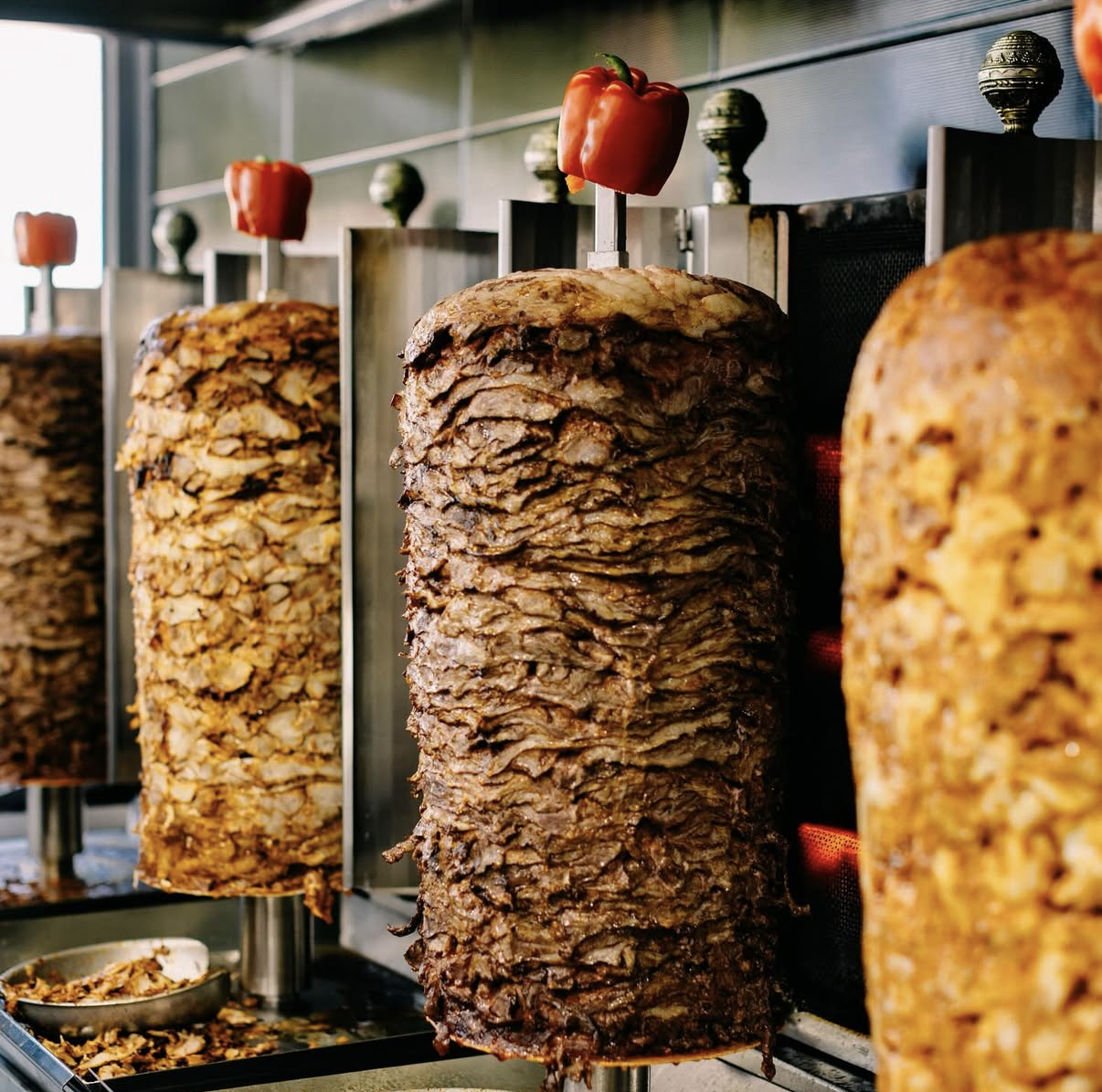 Stacks of seasoned meat on vertical rotisseries, with red peppers on top, in a restaurant or street food setting.