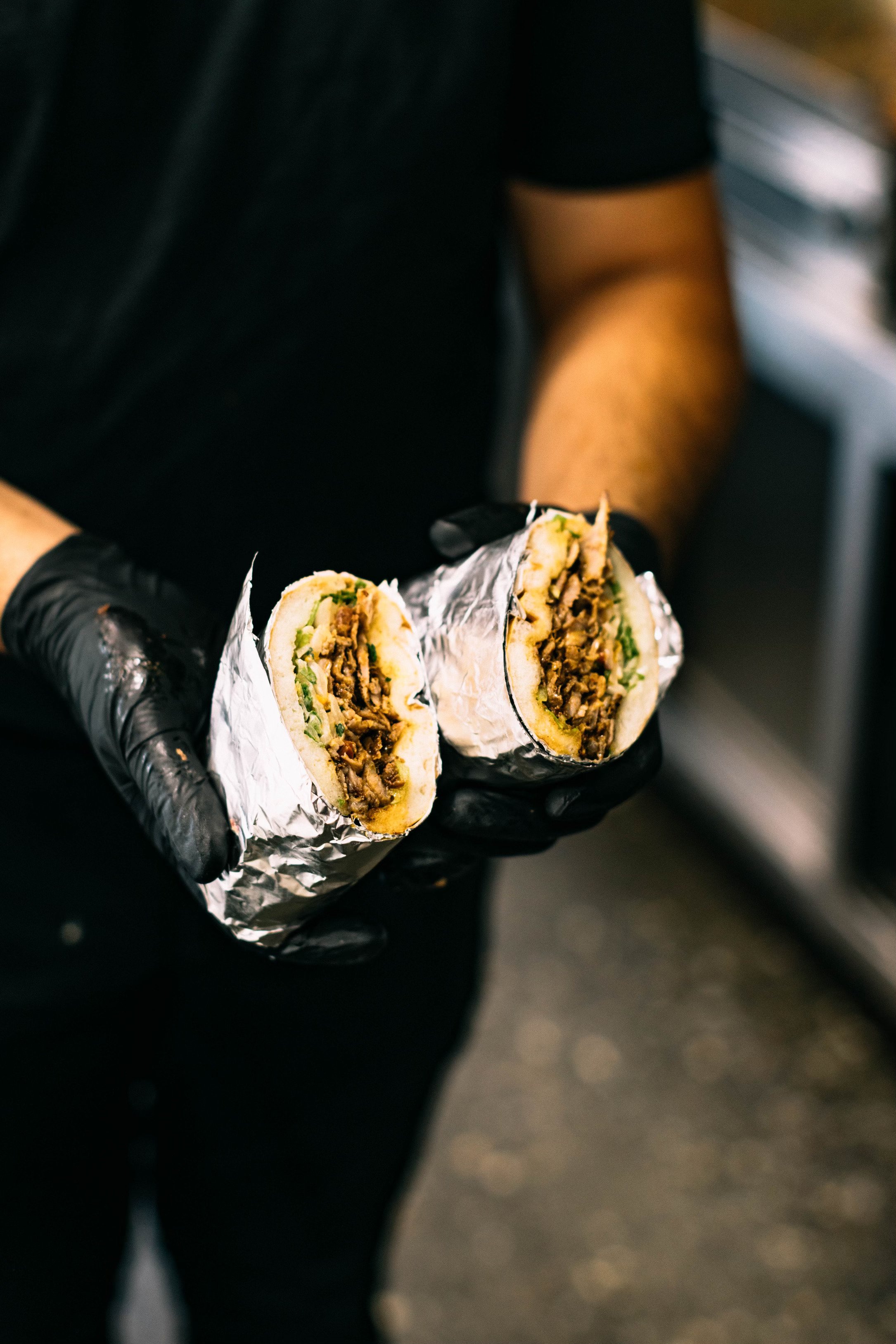 A person wearing black gloves holding a sliced burrito wrapped in aluminum foil.