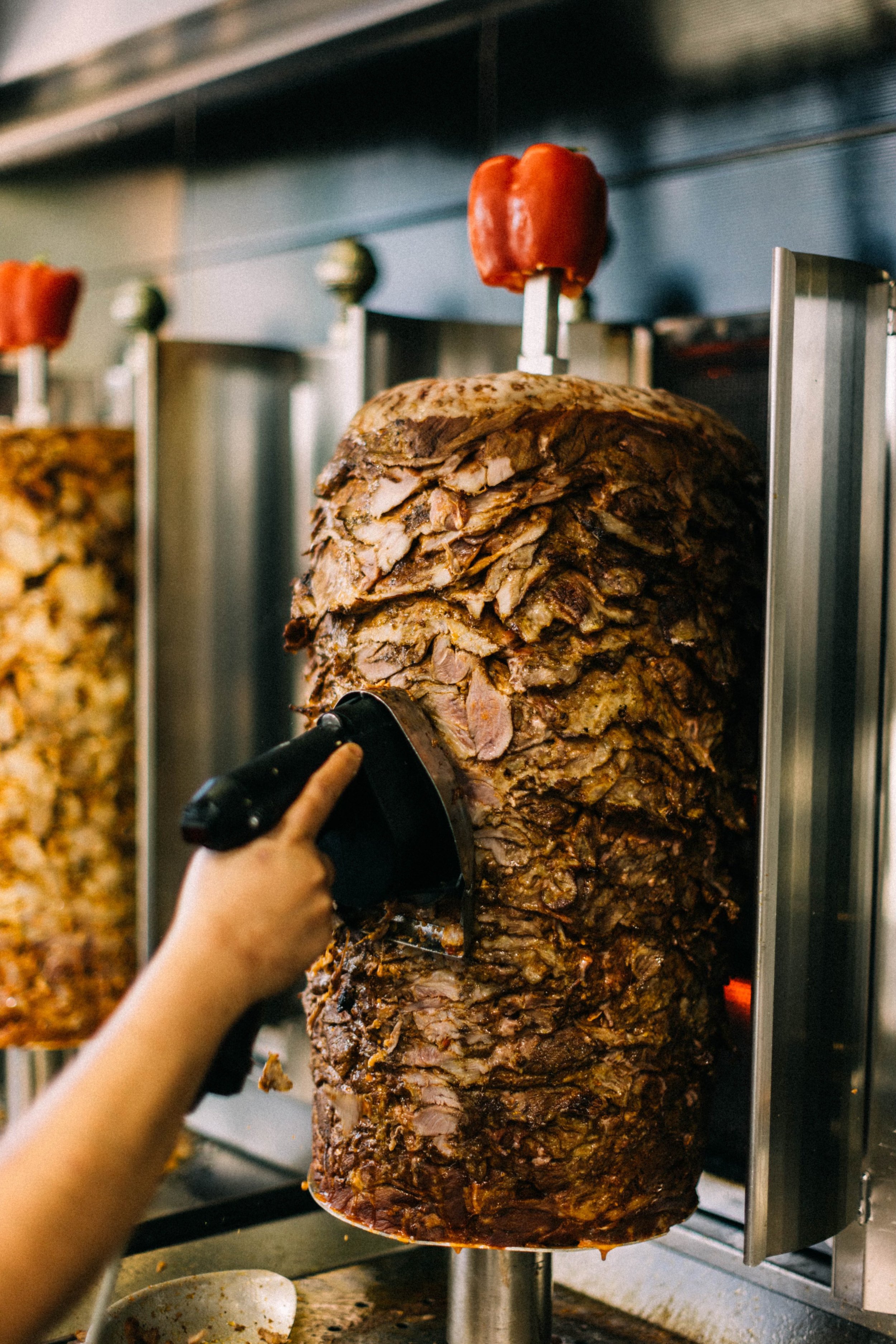 Person carving meat from a large vertical spit machine with red peppers on top.