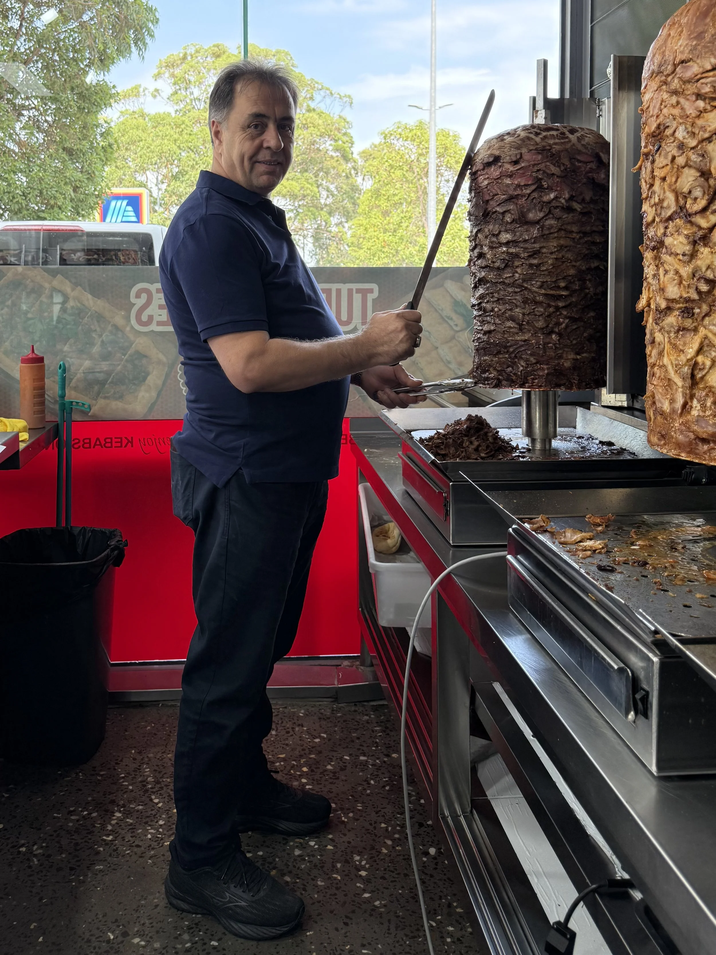 A man in a dark blue shirt and black shoes stands near a vertical rotisserie, slicing meat for shawarma or similar dish inside a food stand with a red counter and utensils behind him. Large portions of cooked meat are spinning on the rotisserie.