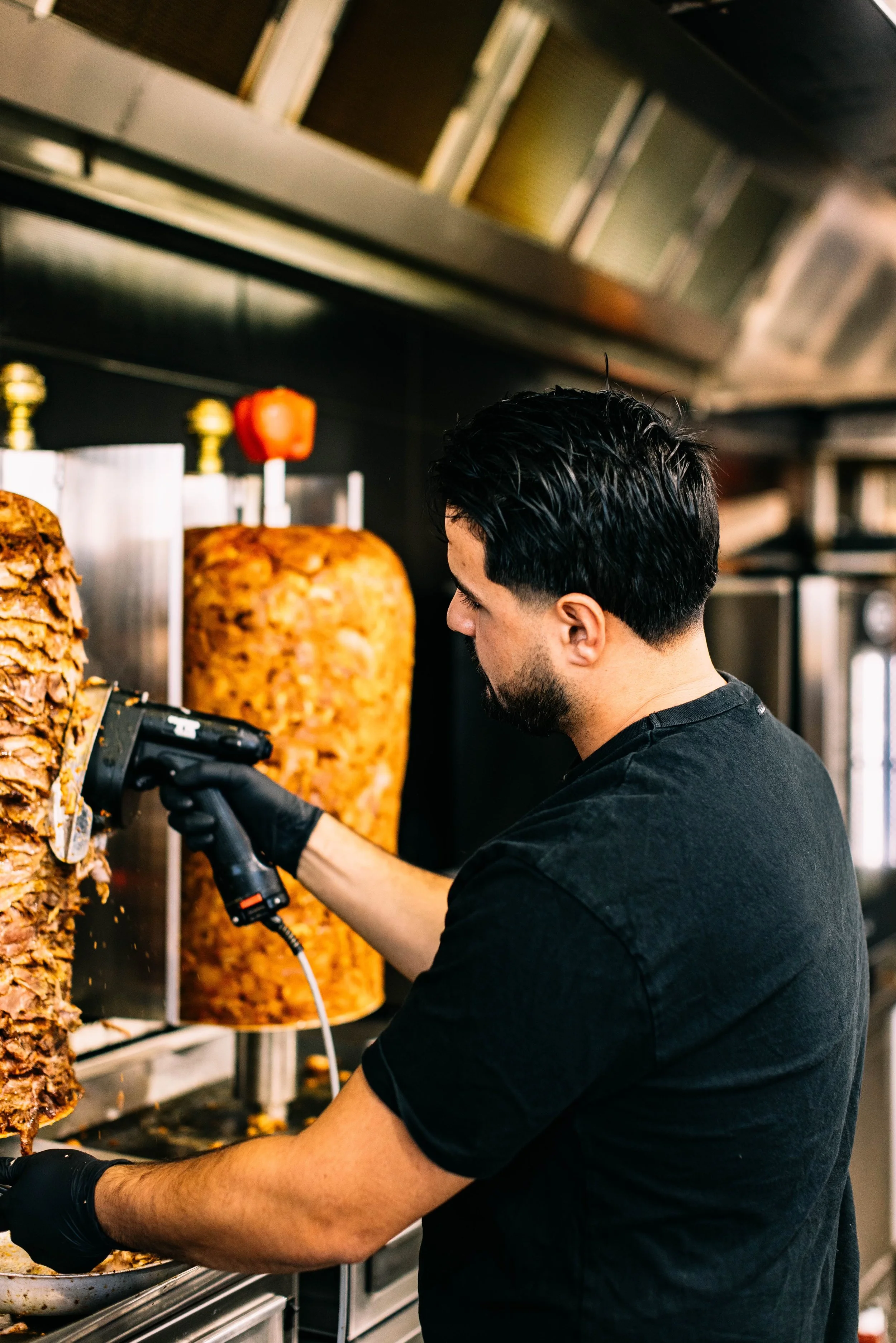 A man with black hair and a beard wearing a black t-shirt and black gloves, slicing roasted meat with an electric knife inside a restaurant kitchen.