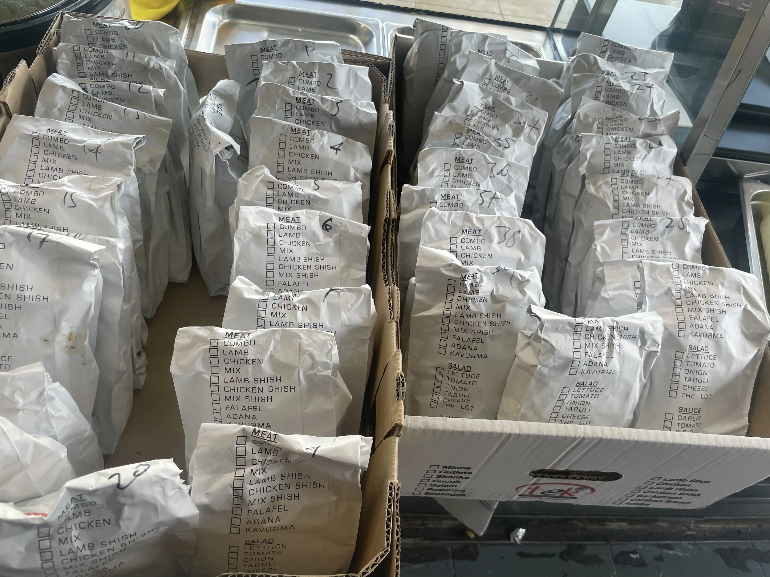 Several paper bags with handwritten numbers and checklists of meat and salad options, placed in a cardboard box.