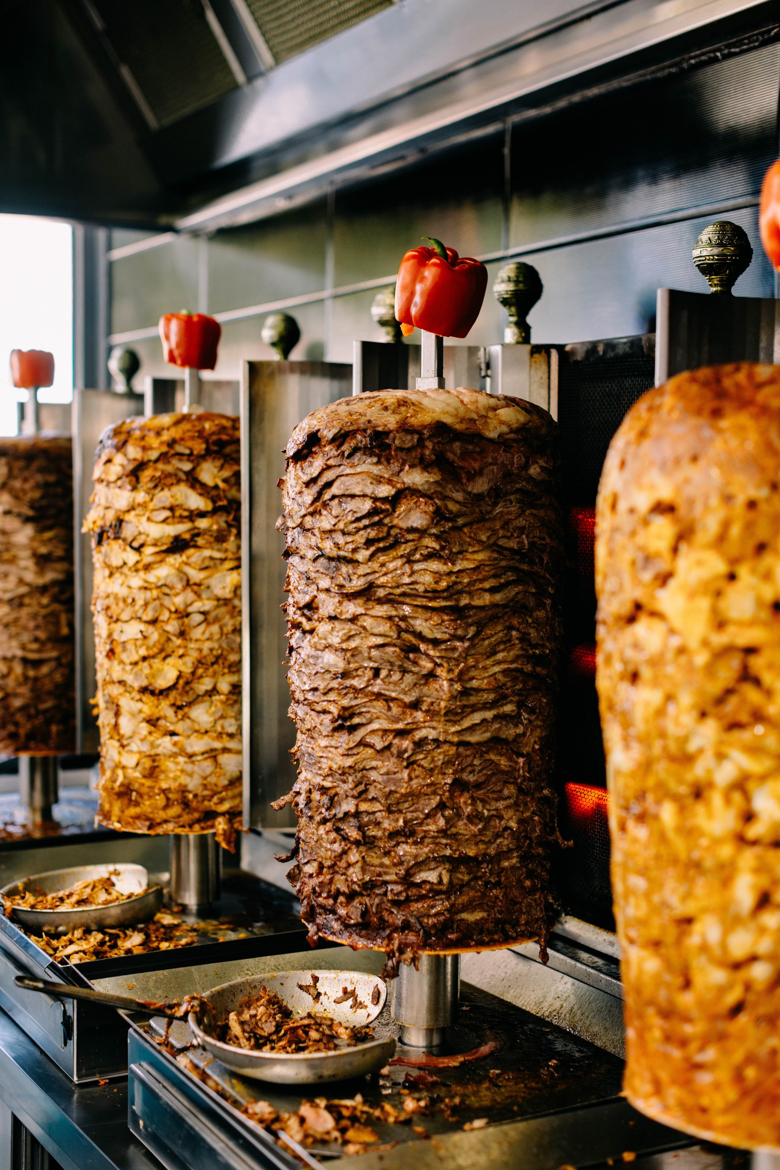 Multiple vertical spits of seasoned meat cooking on a vertical rotisserie, with a tray of cooked meat slices below.