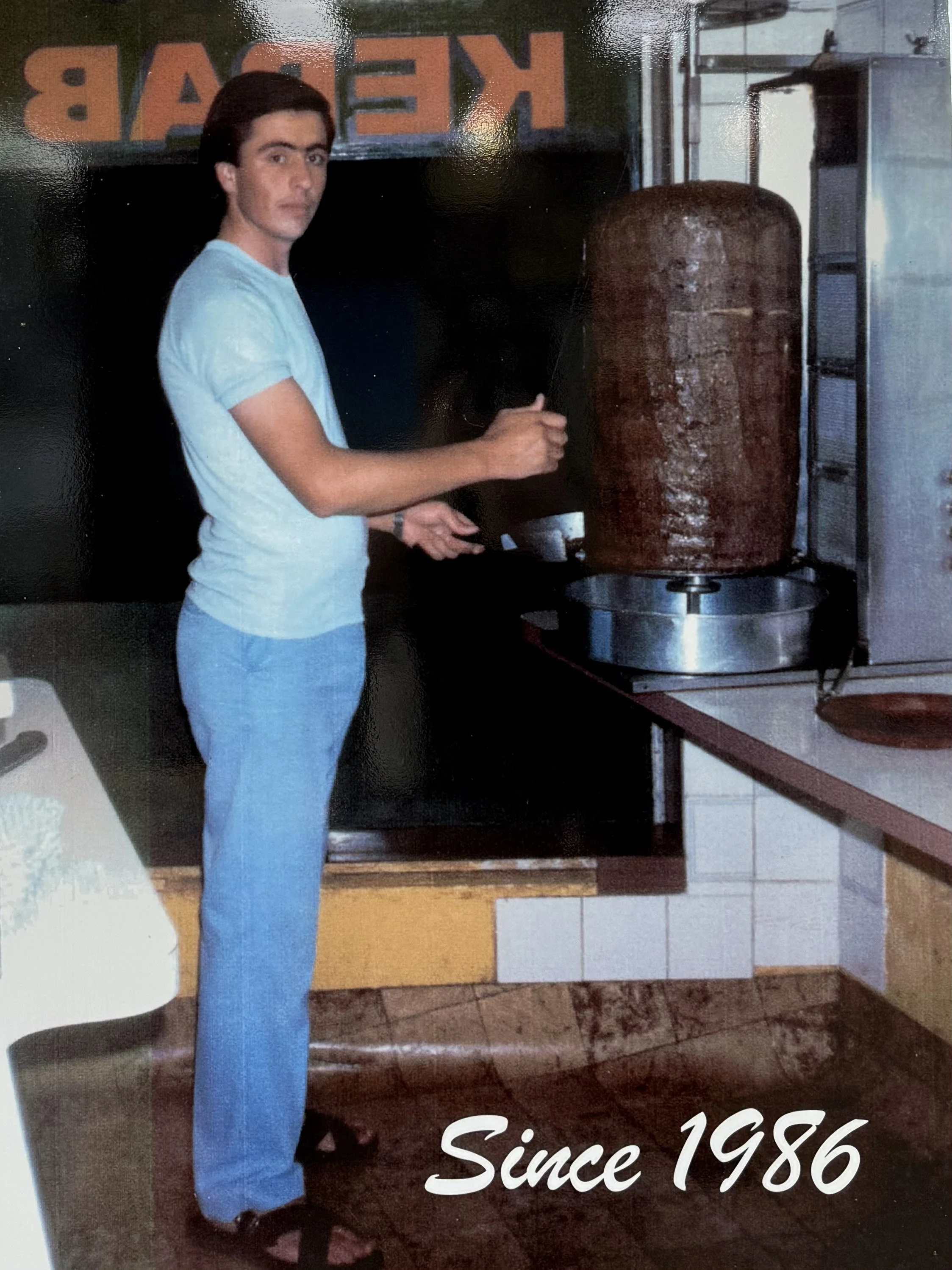 A young man with dark hair, wearing a light blue t-shirt, light blue pants, and slippers, standing in a kitchen and preparing food on a vertical rotisserie of meat.
