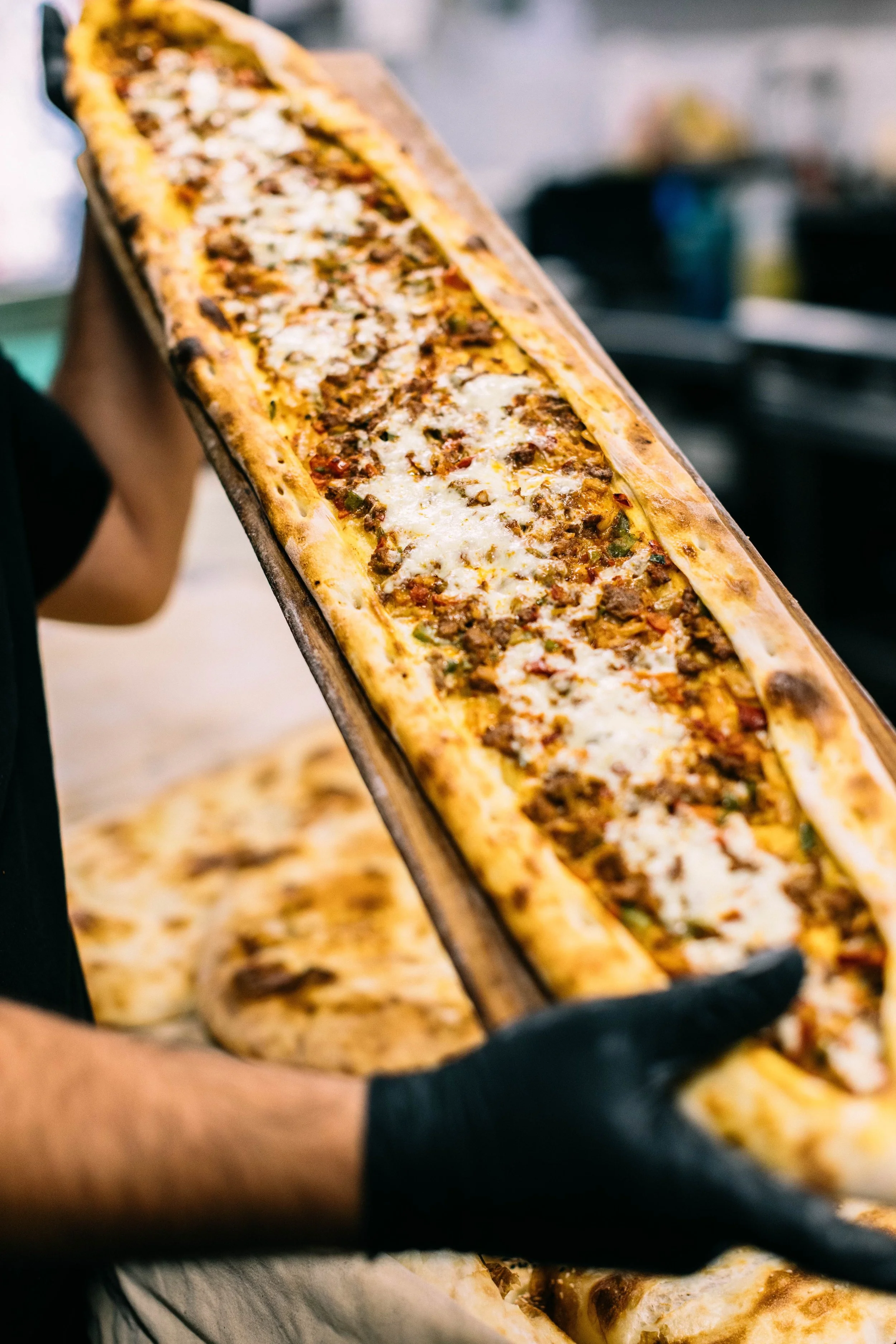 A person holding a long rectangular pizza with various toppings, including cheese, meat, and vegetables, on a wooden pizza peel.