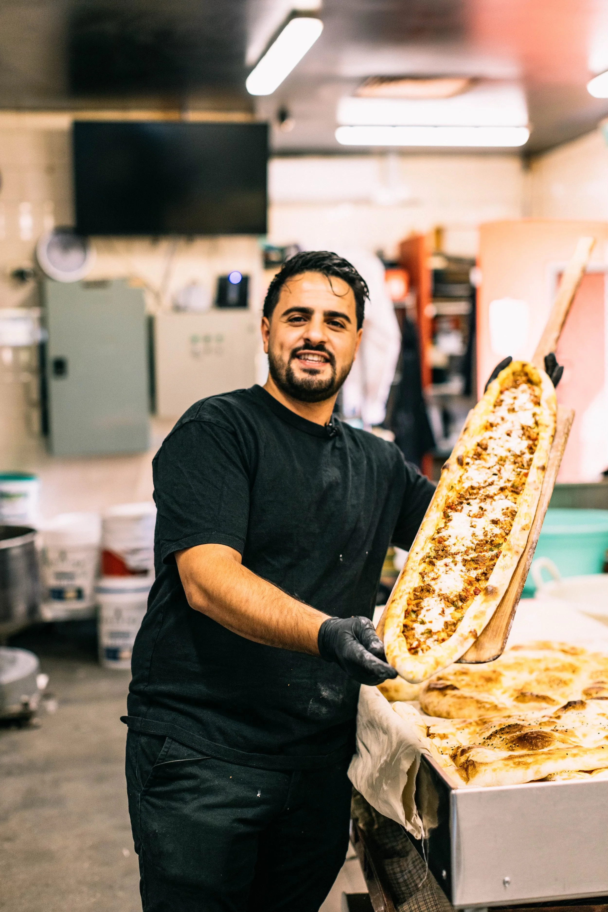 A man in a black shirt and gloves holding a large pizza with various toppings.