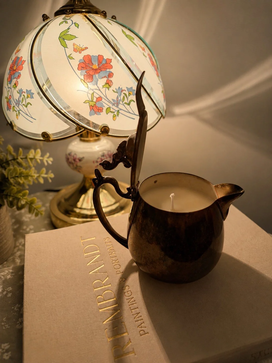 A vintage table lamp with floral glass shade, a dark ceramic teapot-shaped candle holder, a paperback 'The Hand Painting & Portraits' book, and a potted plant on a lace tablecloth.