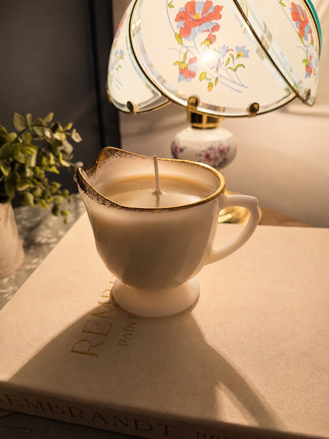 A lit candle in a vintage ceramic pitcher-shaped holder with a small handle, placed on a book titled "Rembrandt." In the background, an ornate floral table lamp with a curved glass shade and a porcelain base, along with a small potted plant, are visi