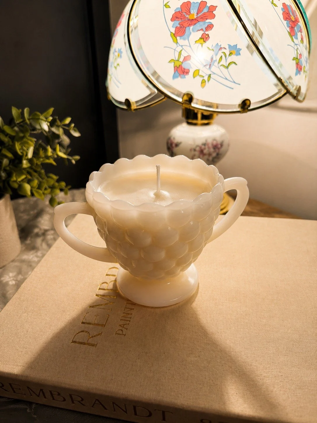 A white grape-themed candle holder with a candle inside, placed on a beige box with text, next to a potted plant, with a floral-patterned table lamp in the background.