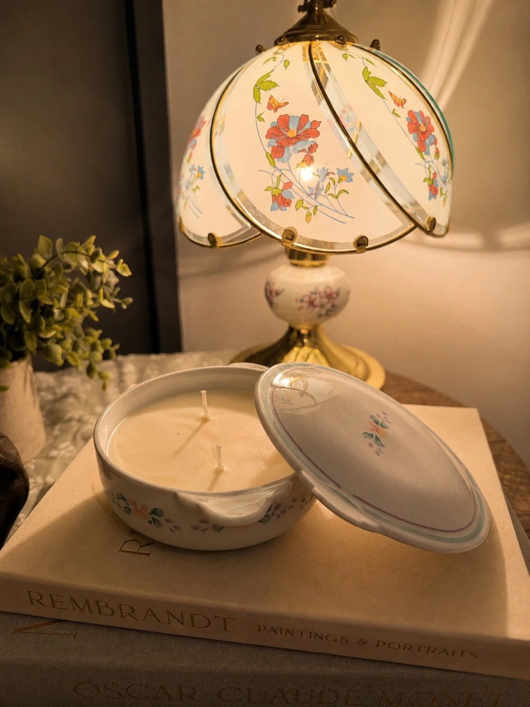 A decorative table lamp with floral designs, a teacup-shaped candle holder, and a book titled 'Rembrandt Paintings & Portraits' on a table, with a small plant nearby.